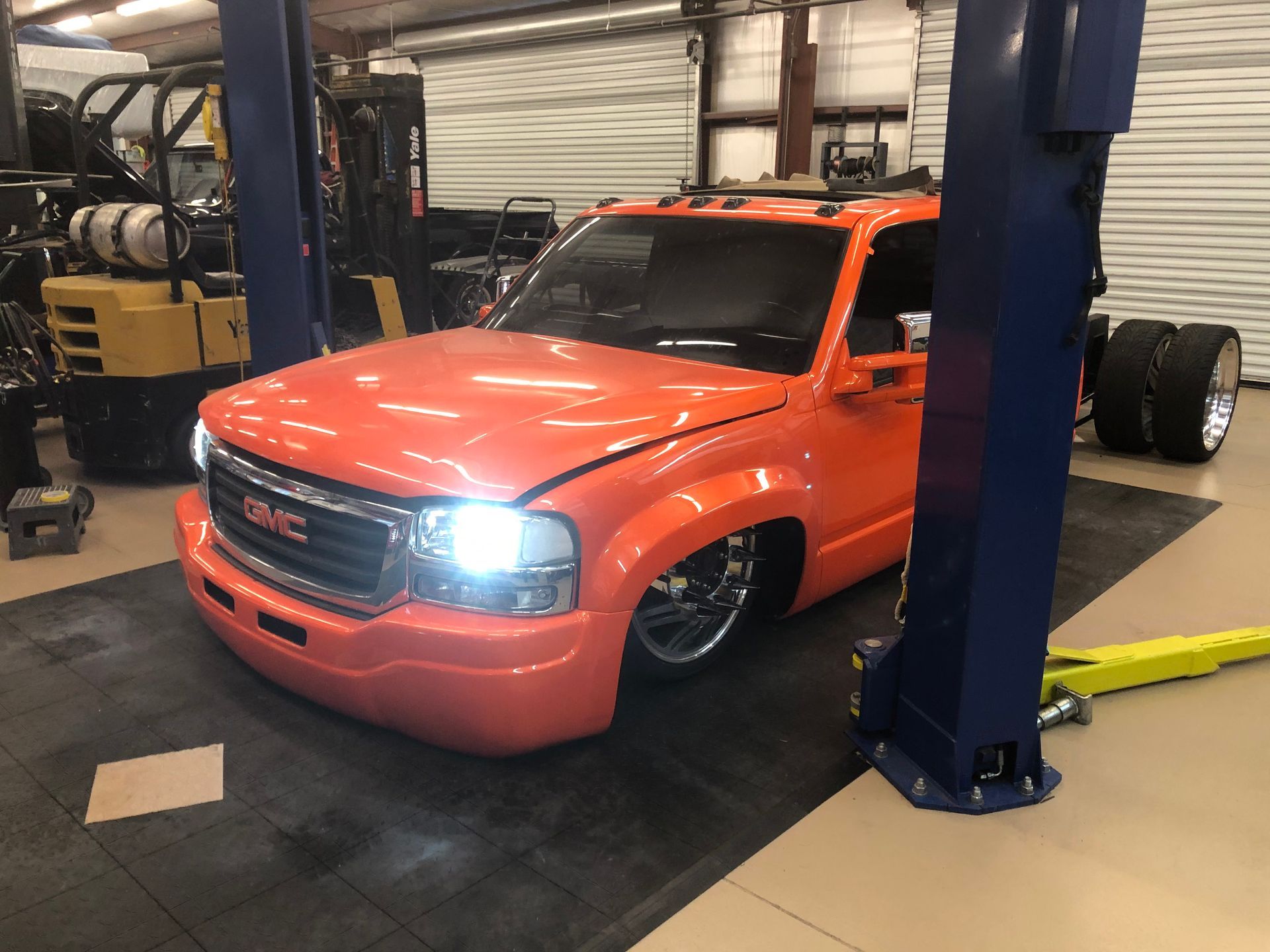 A bright orange lowered GMC pickup truck parked inside a workshop between two metal car lift pillars.
