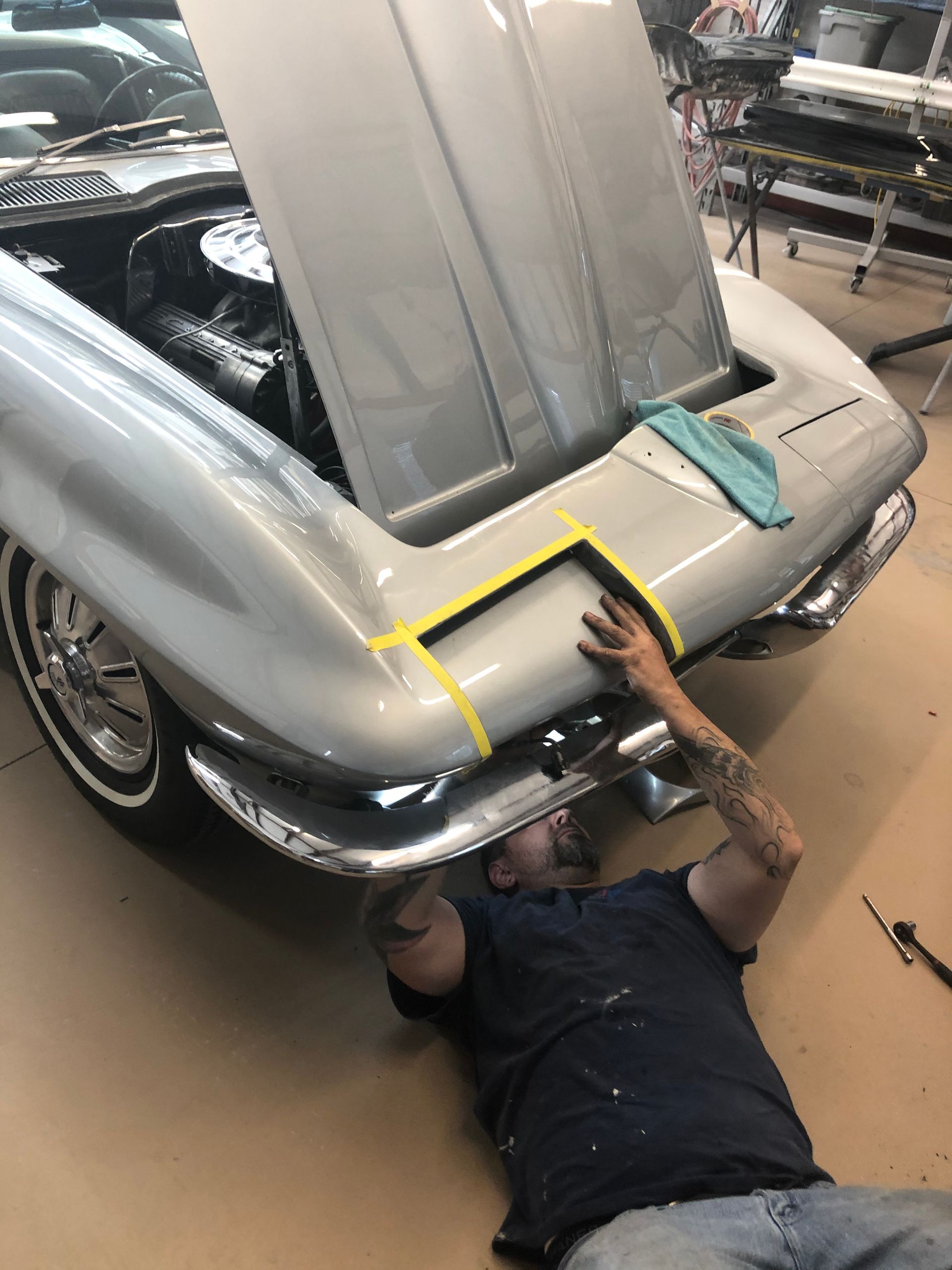 A mechanic works on the front light assembly of a silver vintage Corvette in an auto shop.