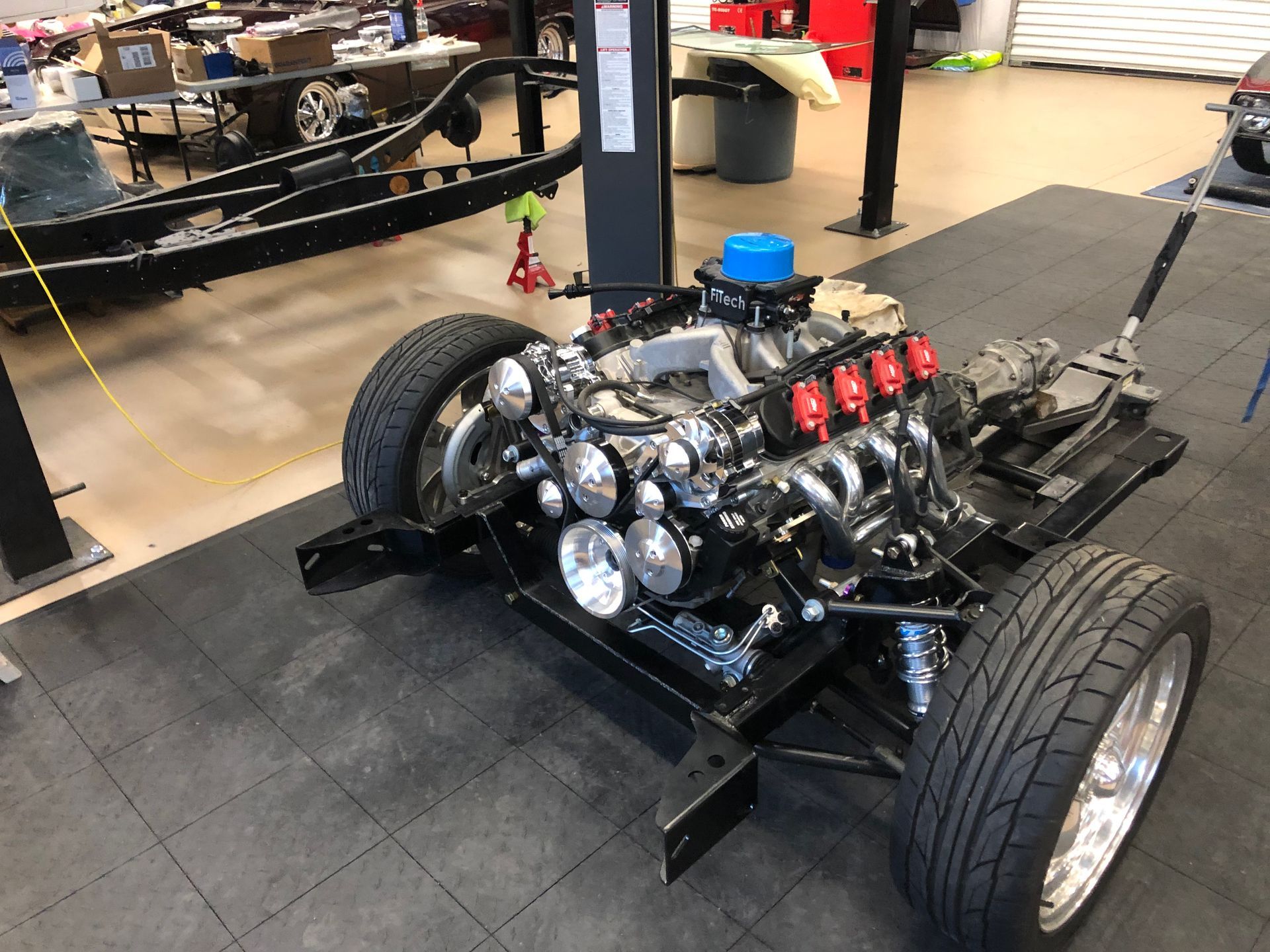 A partially assembled vehicle chassis with an engine and front wheels resting on a tiled floor in a garage.