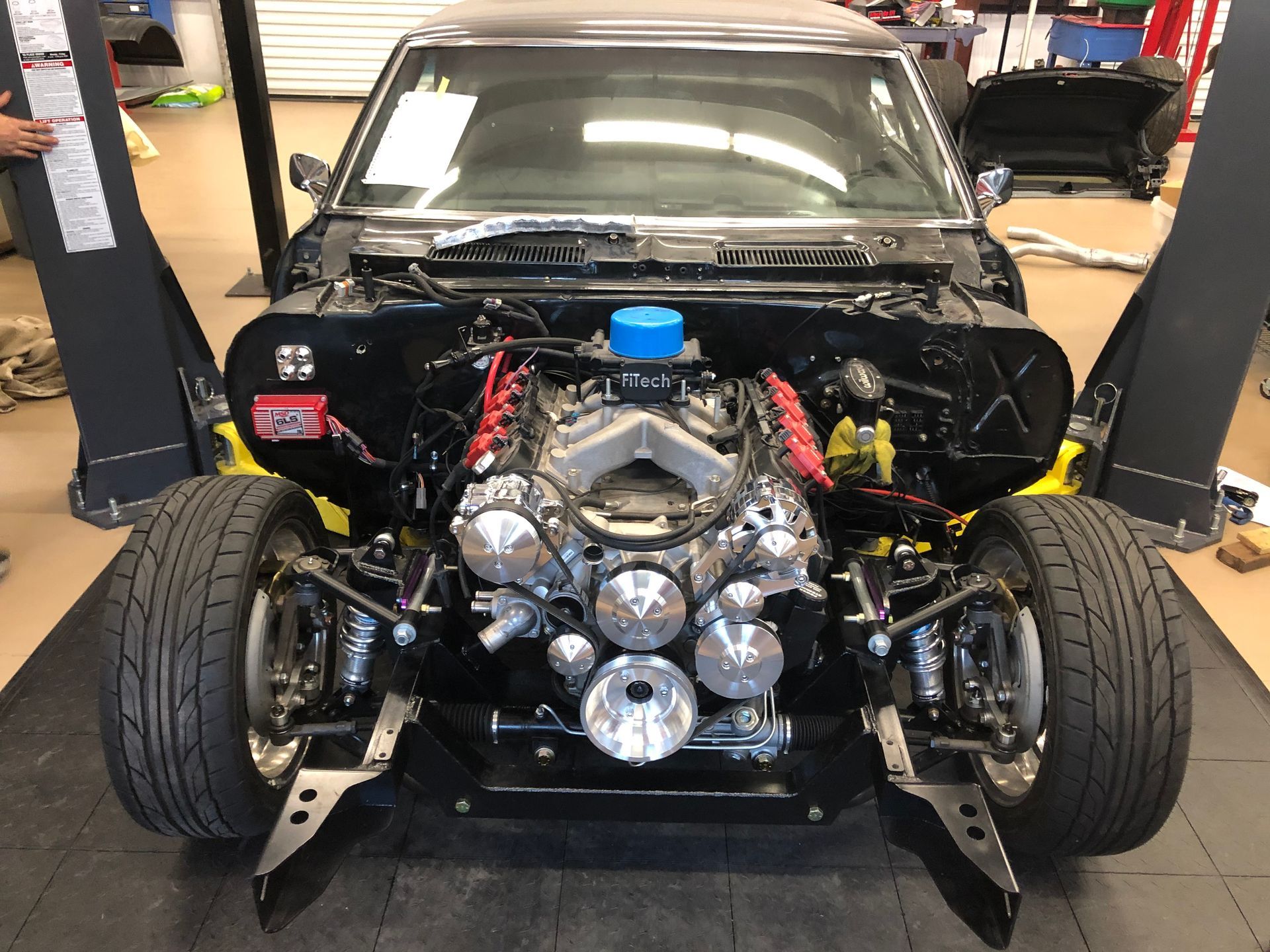 Front view of a black muscle car chassis in a garage, featuring a highly detailed chrome engine and exposed suspension.