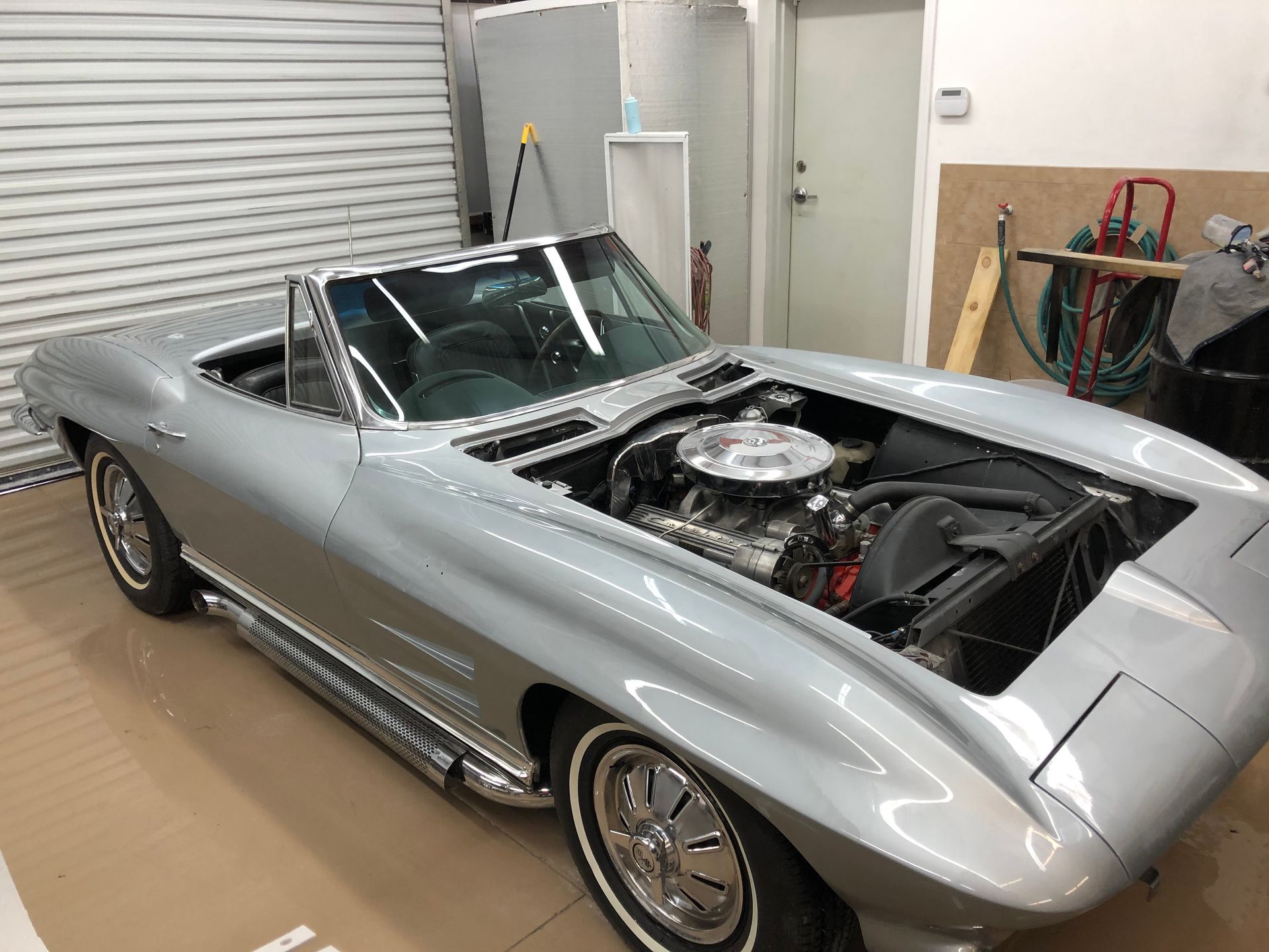 A silver 1963 Chevrolet Corvette convertible with its hood removed, parked in a garage.