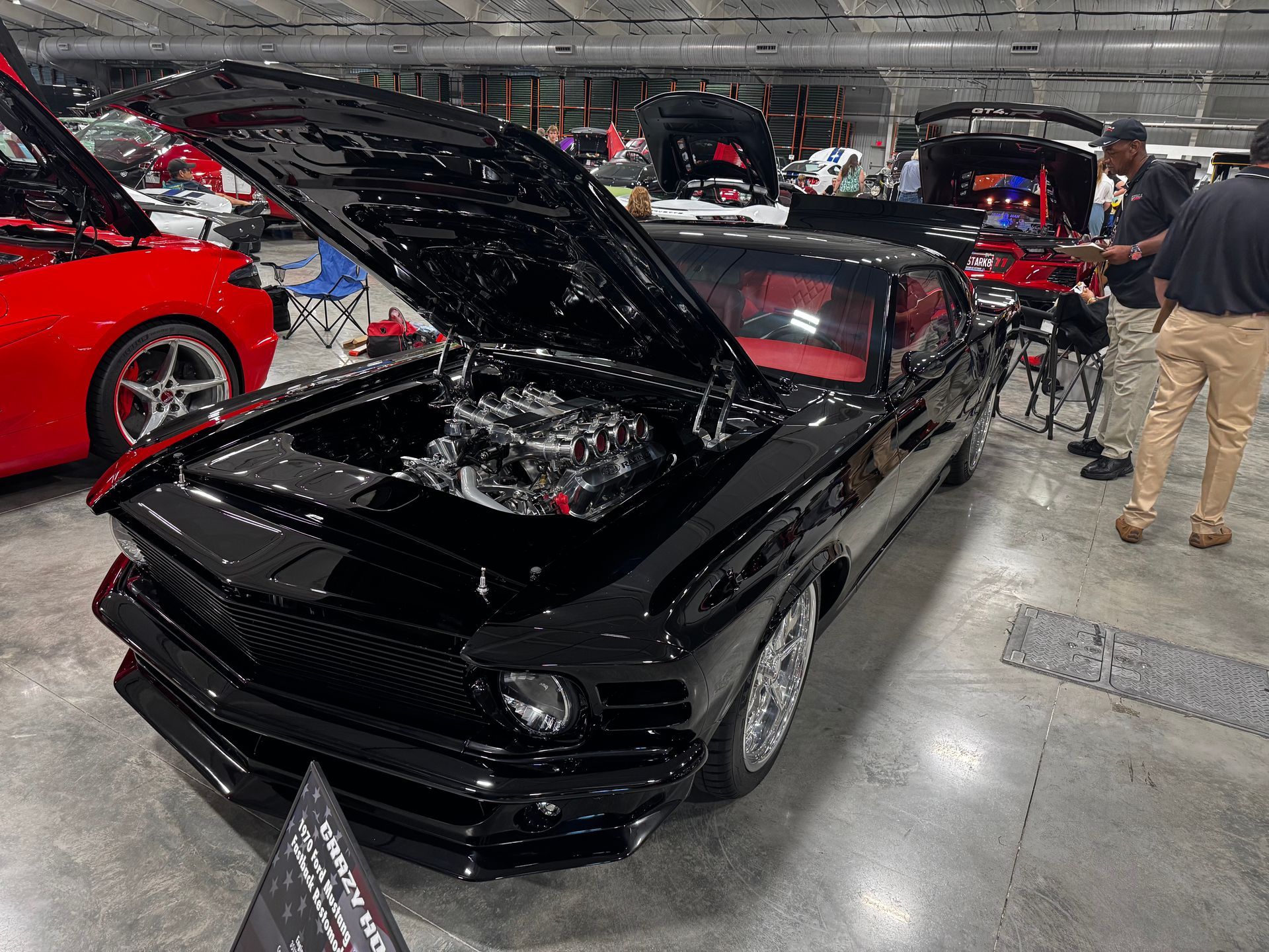 A glossy black 1969 Ford Mustang with its hood open, displayed on a concrete floor at an indoor car show.