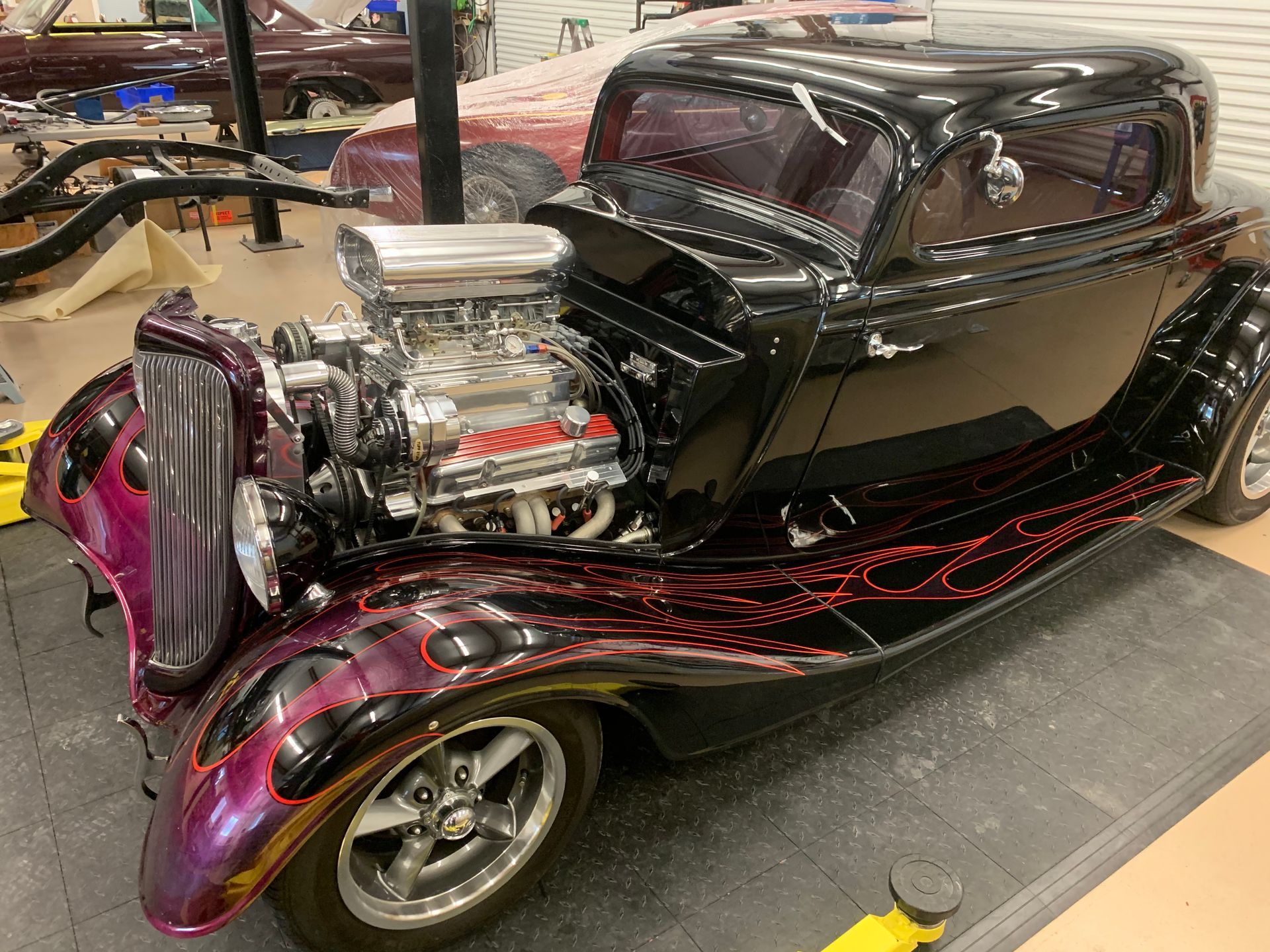A black hot rod with exposed chrome engine and custom purple flame paint on fenders sits inside a garage.