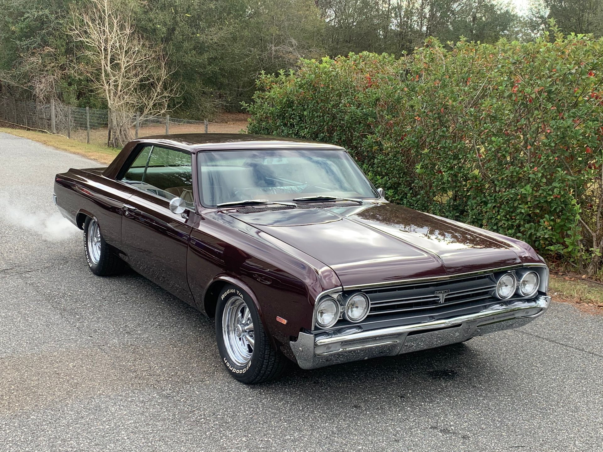A dark maroon vintage muscle car parked on a gravel road in front of green bushes.