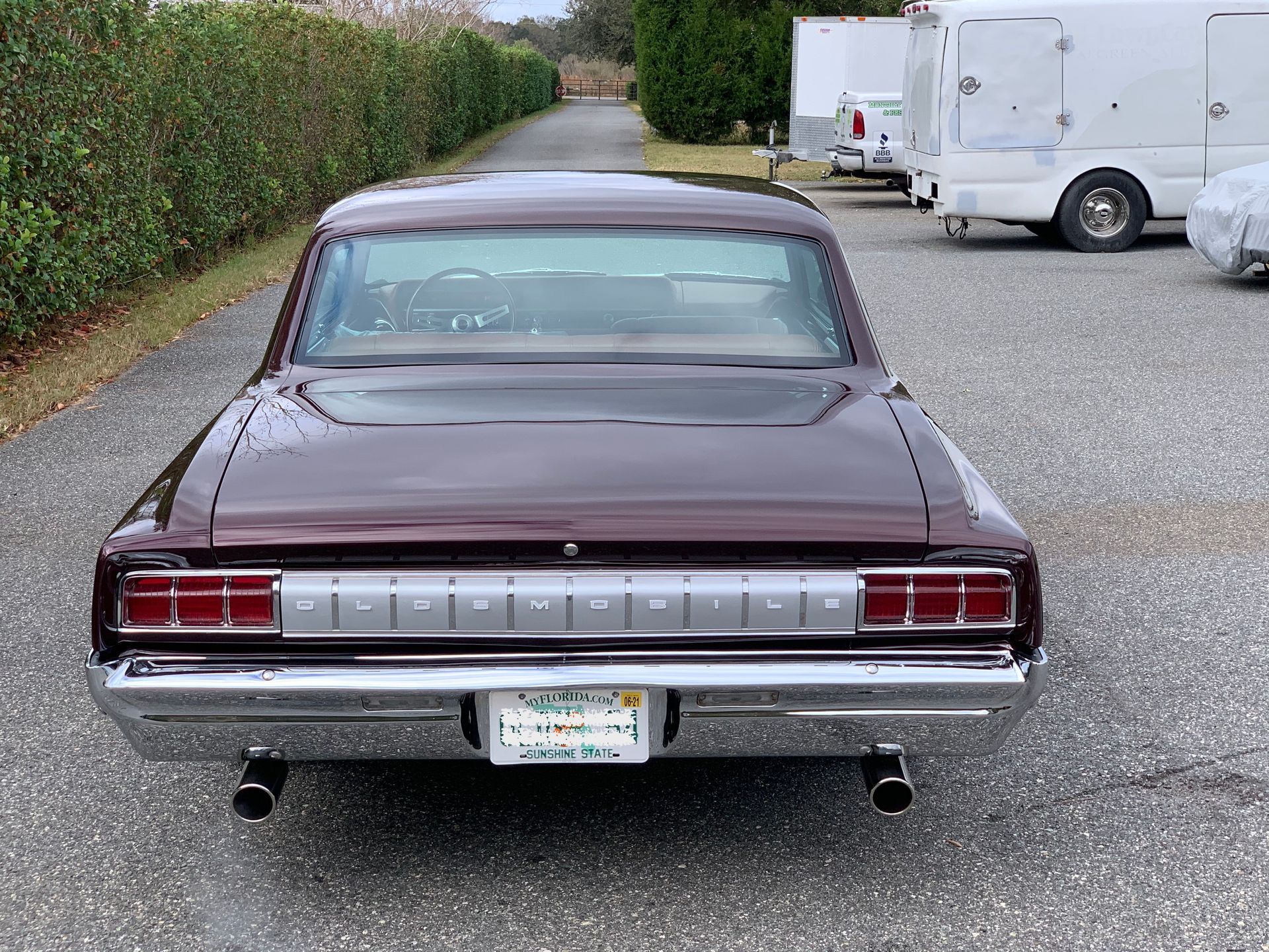 A rear view of a dark maroon 1964 Oldsmobile parked on a gravel surface next to a white trailer.