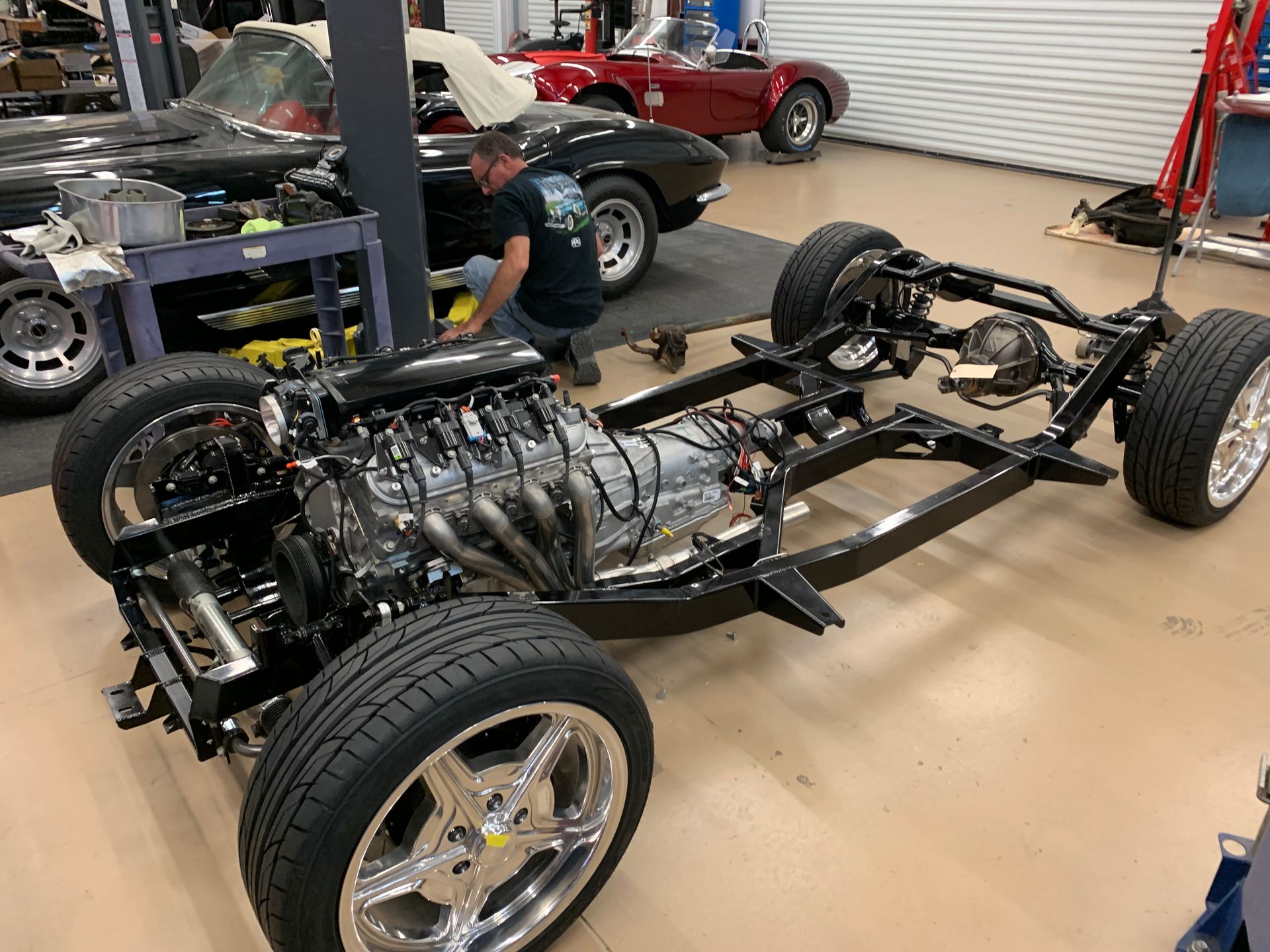 A mechanic works on a classic car chassis with an installed engine, in an auto shop with another car in the background.