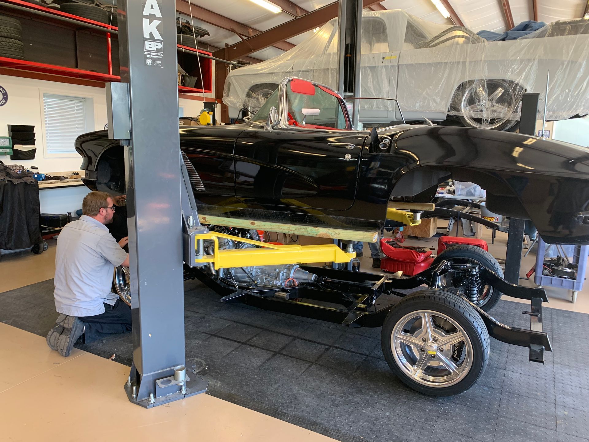 A mechanic works on the chassis of a black convertible car elevated on a yellow hydraulic lift in a garage.