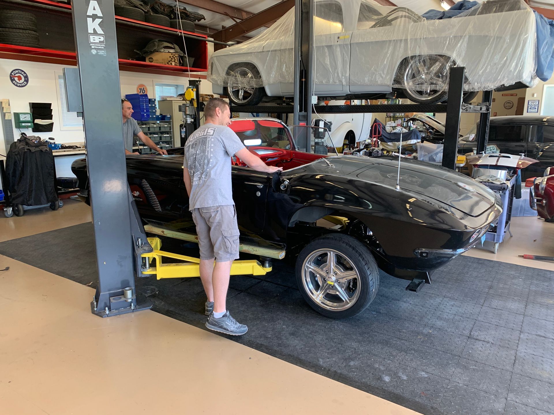 A person in a gray shirt stands next to a black classic car inside a garage with a lift holding another vehicle.