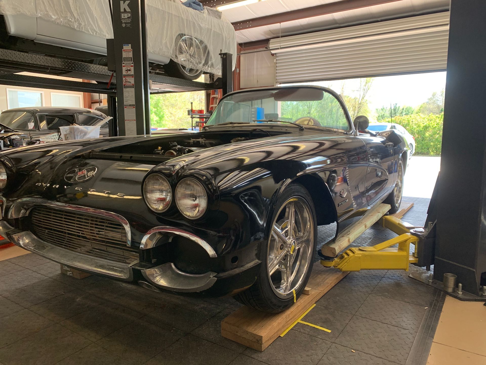 A black classic Chevrolet Corvette convertible lifted on a yellow hydraulic auto repair lift in a garage.