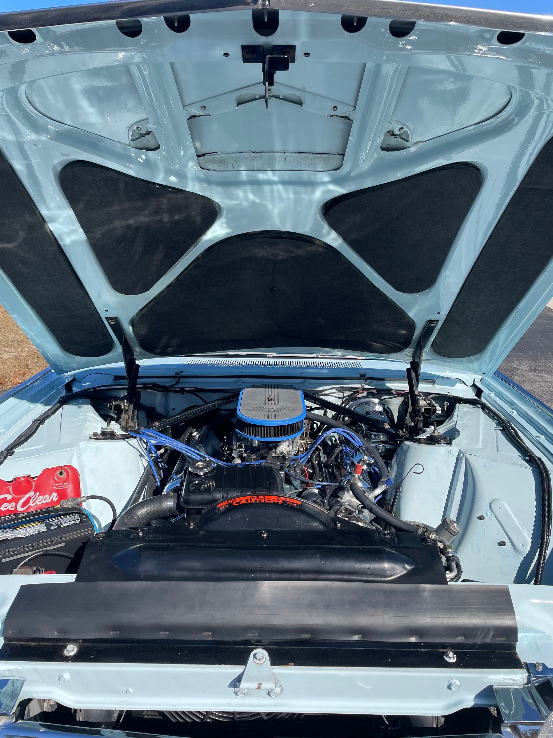 The open hood of a light blue classic car reveals a detailed engine bay with a red battery and dark cooling fan.