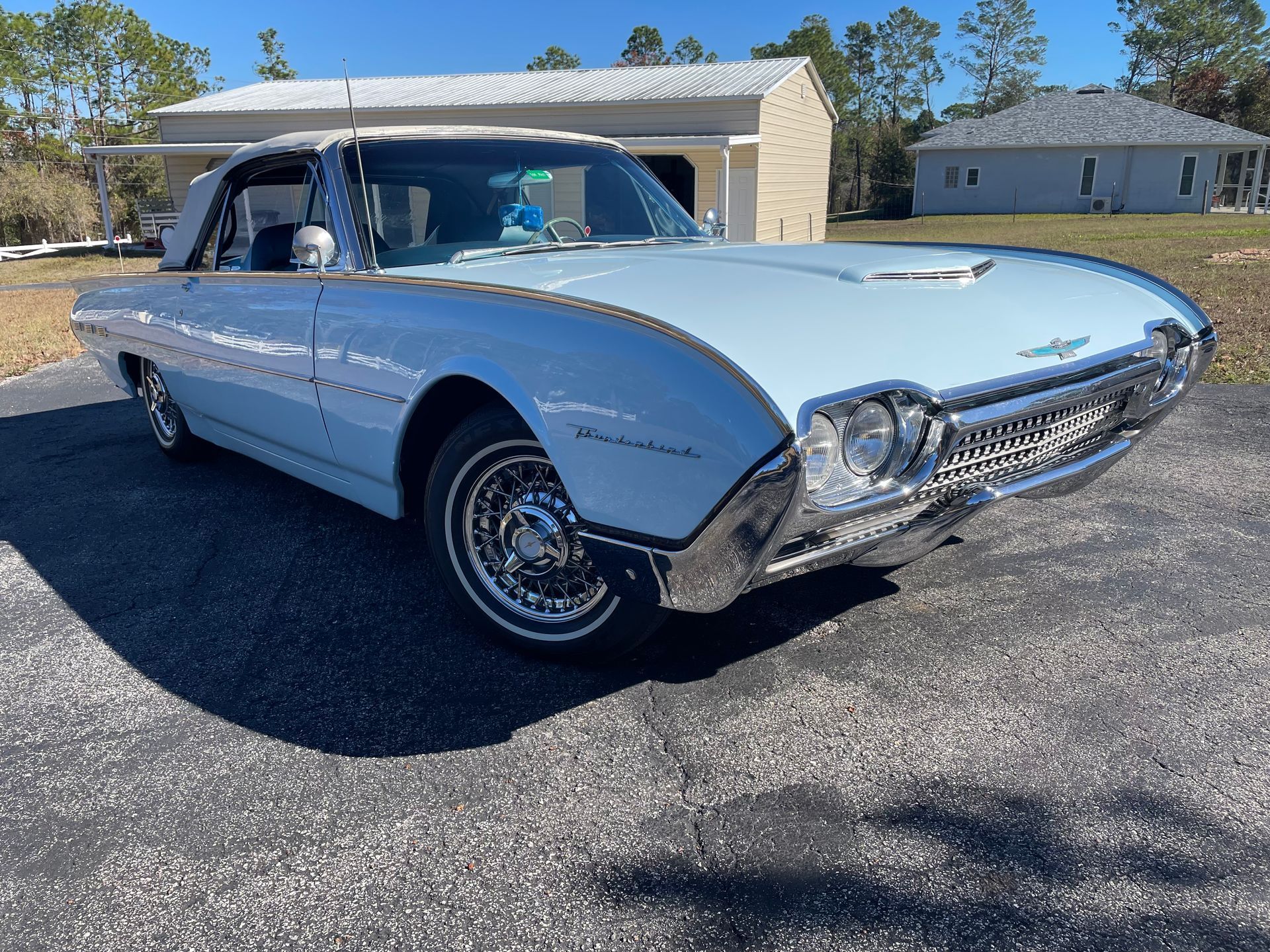 A light blue vintage Ford Thunderbird convertible parked on an asphalt driveway on a sunny day.