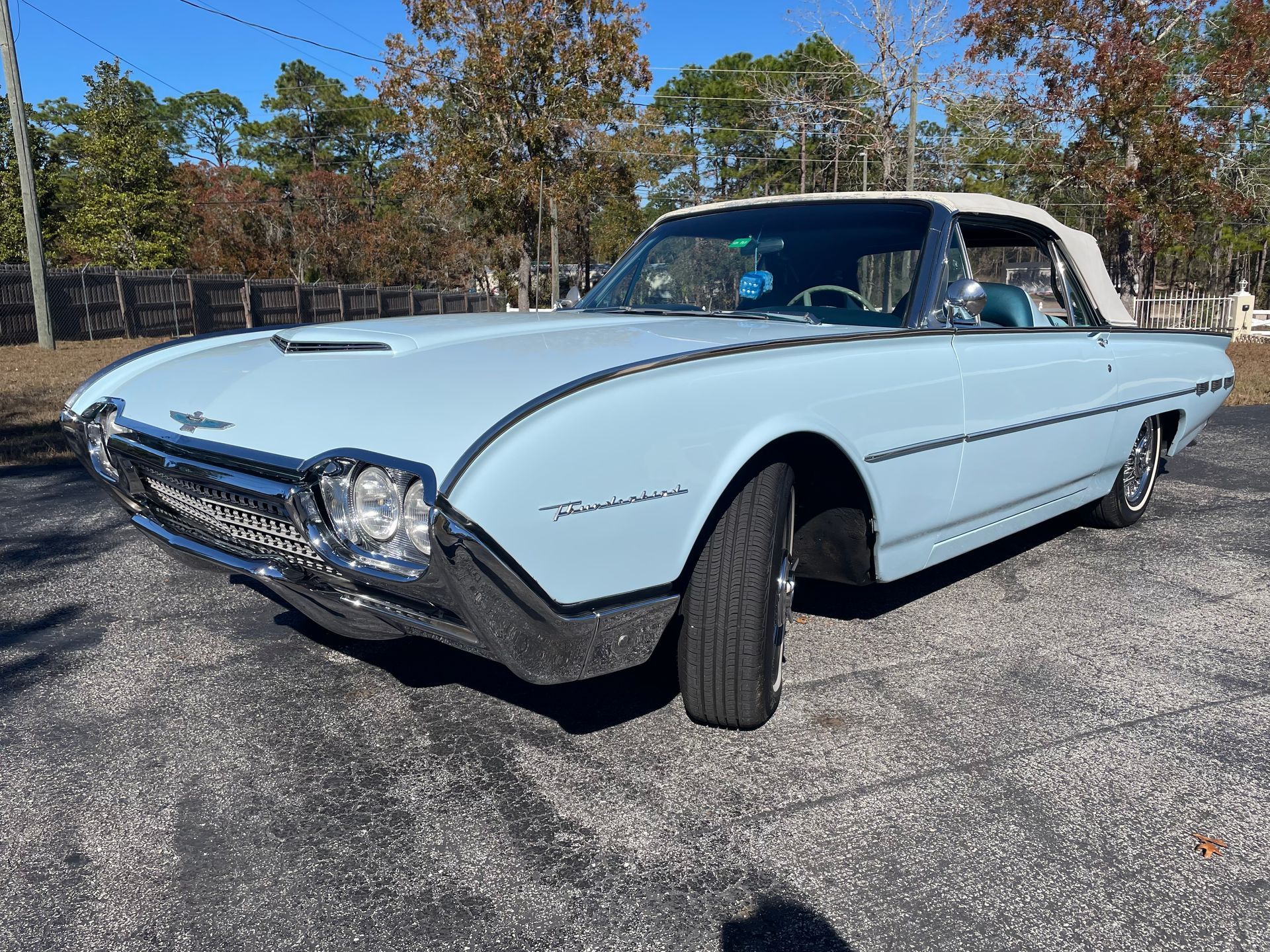 A light blue 1962 Ford Thunderbird convertible parked on an asphalt lot with trees in the background.