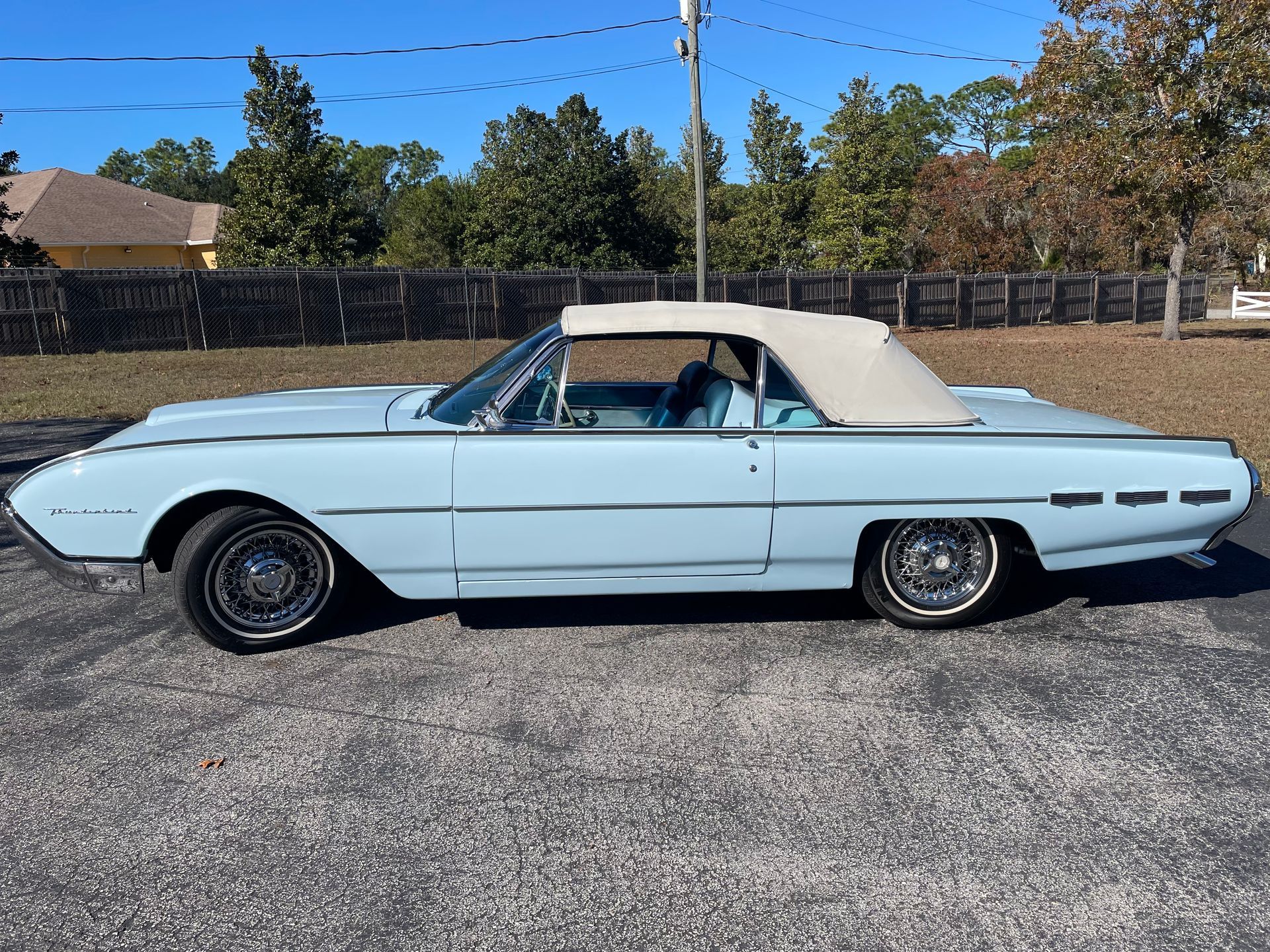 A light blue vintage Ford Thunderbird convertible with a tan soft top parked on an asphalt lot.