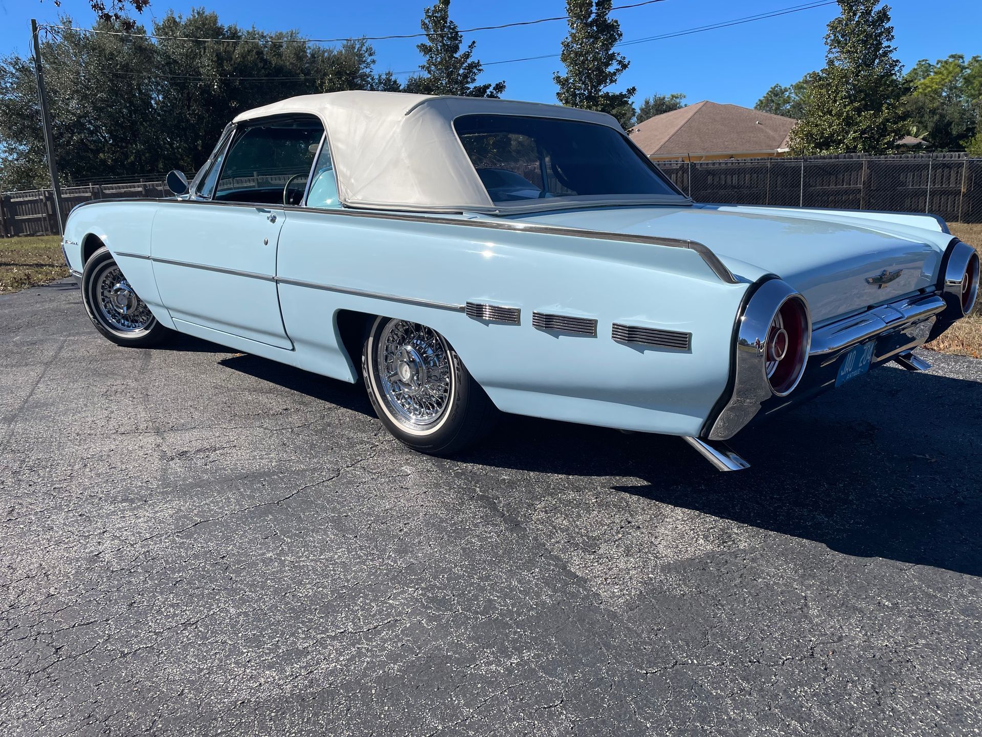 A light blue 1962 Ford Thunderbird convertible with a tan top parked on an asphalt lot under a clear blue sky.