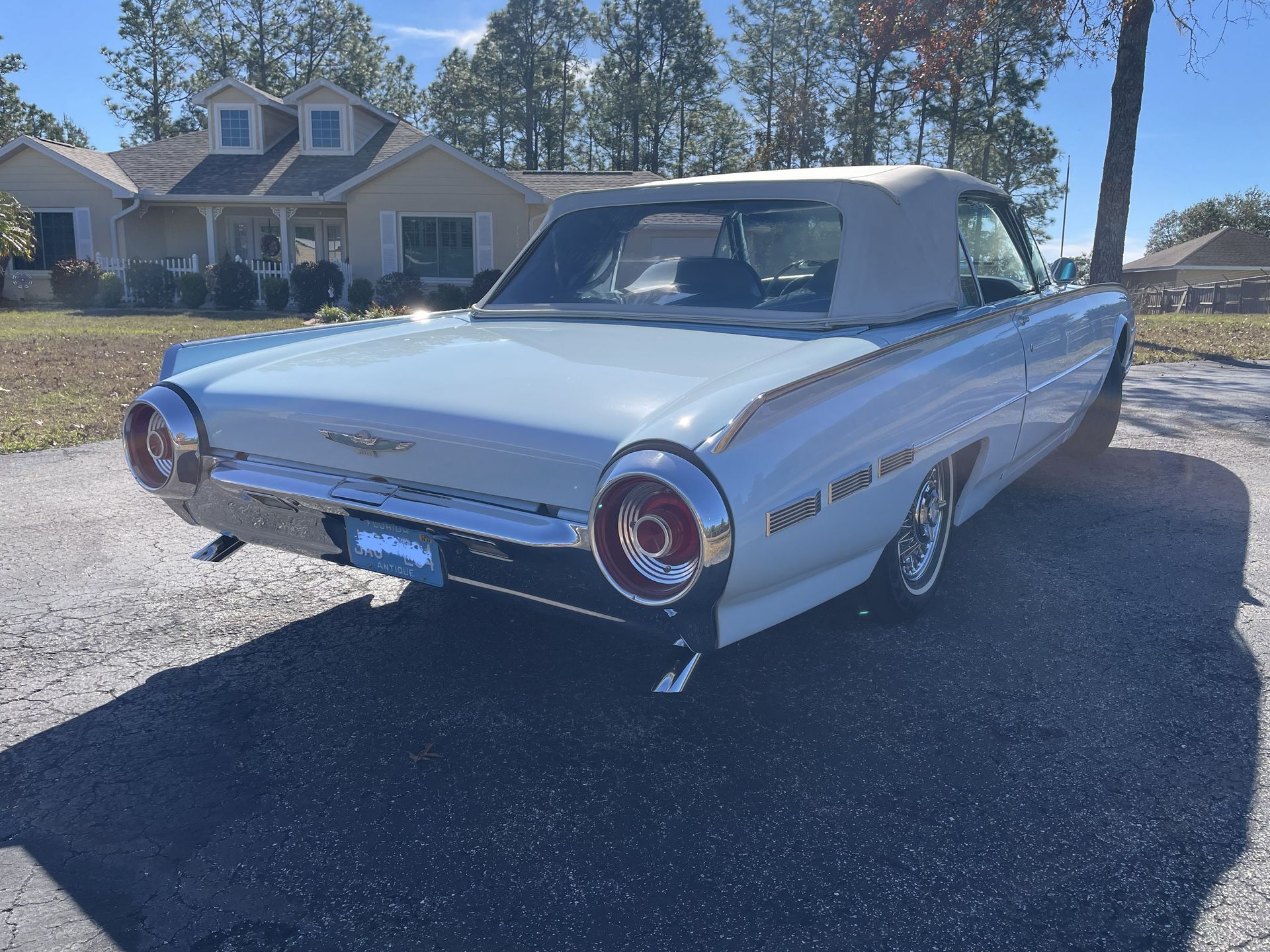 A light blue vintage Ford Thunderbird convertible parked on a gravel driveway in front of a suburban house.