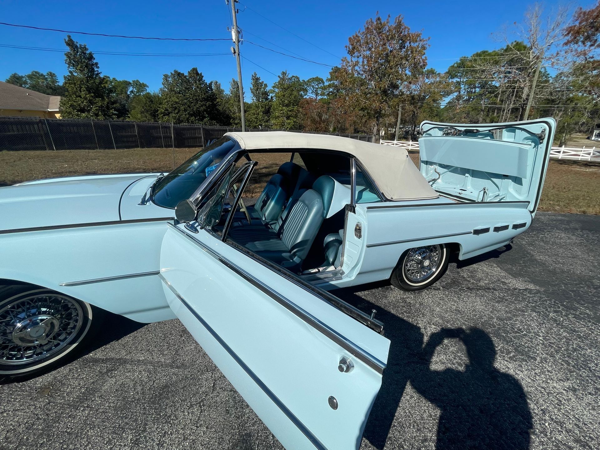 A vintage, light blue convertible car with its door and trunk open, parked on an asphalt surface under a clear blue sky.