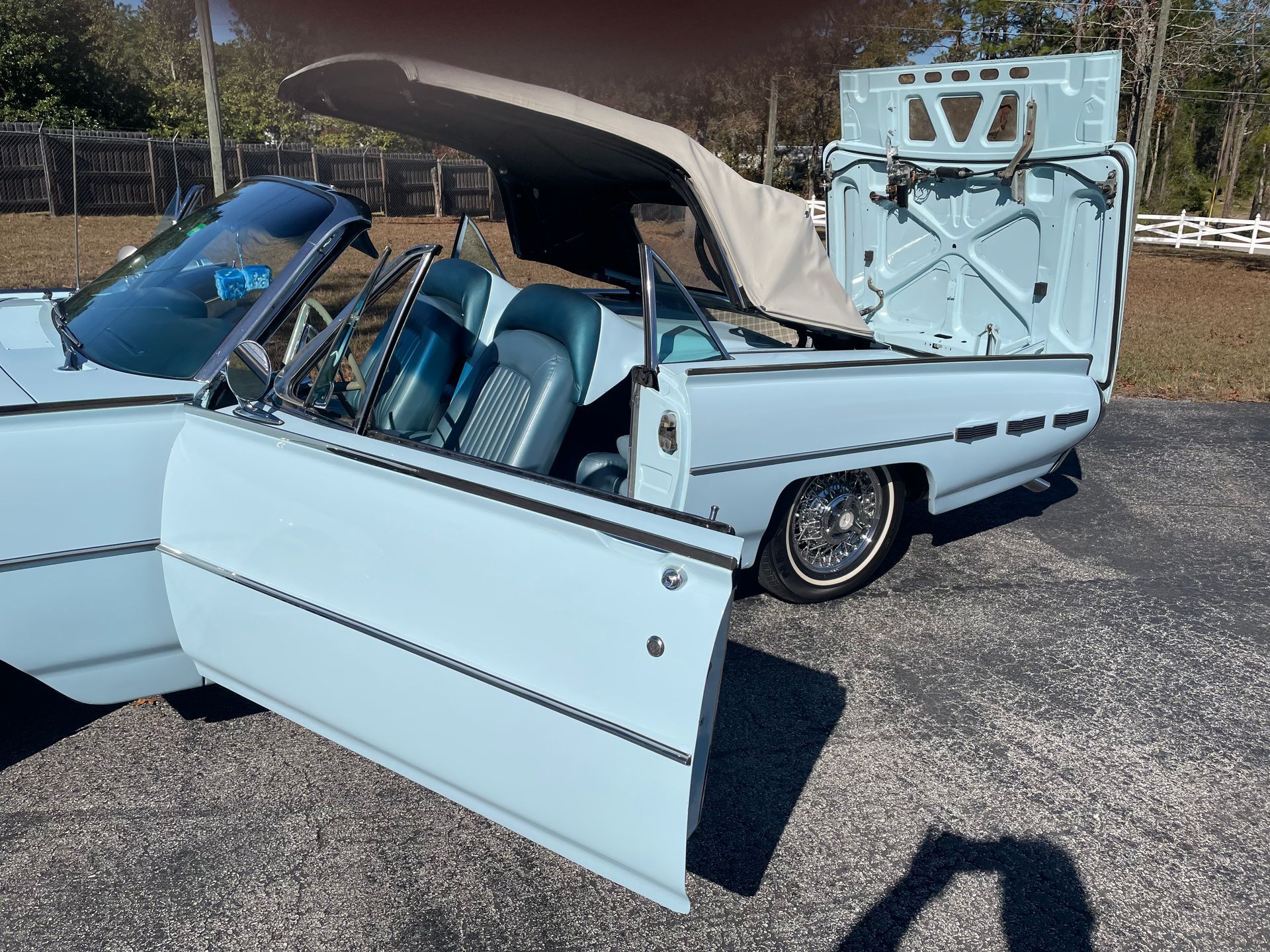 A light blue 1960s Ford Thunderbird convertible with its trunk lid open and the retractable hardtop mid-cycle.