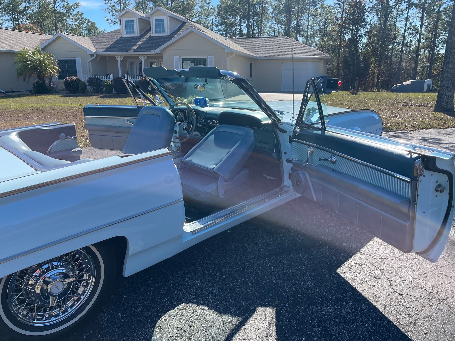 A light blue classic convertible car with chrome wire wheels and an open door parked in front of a house on a sunny day.