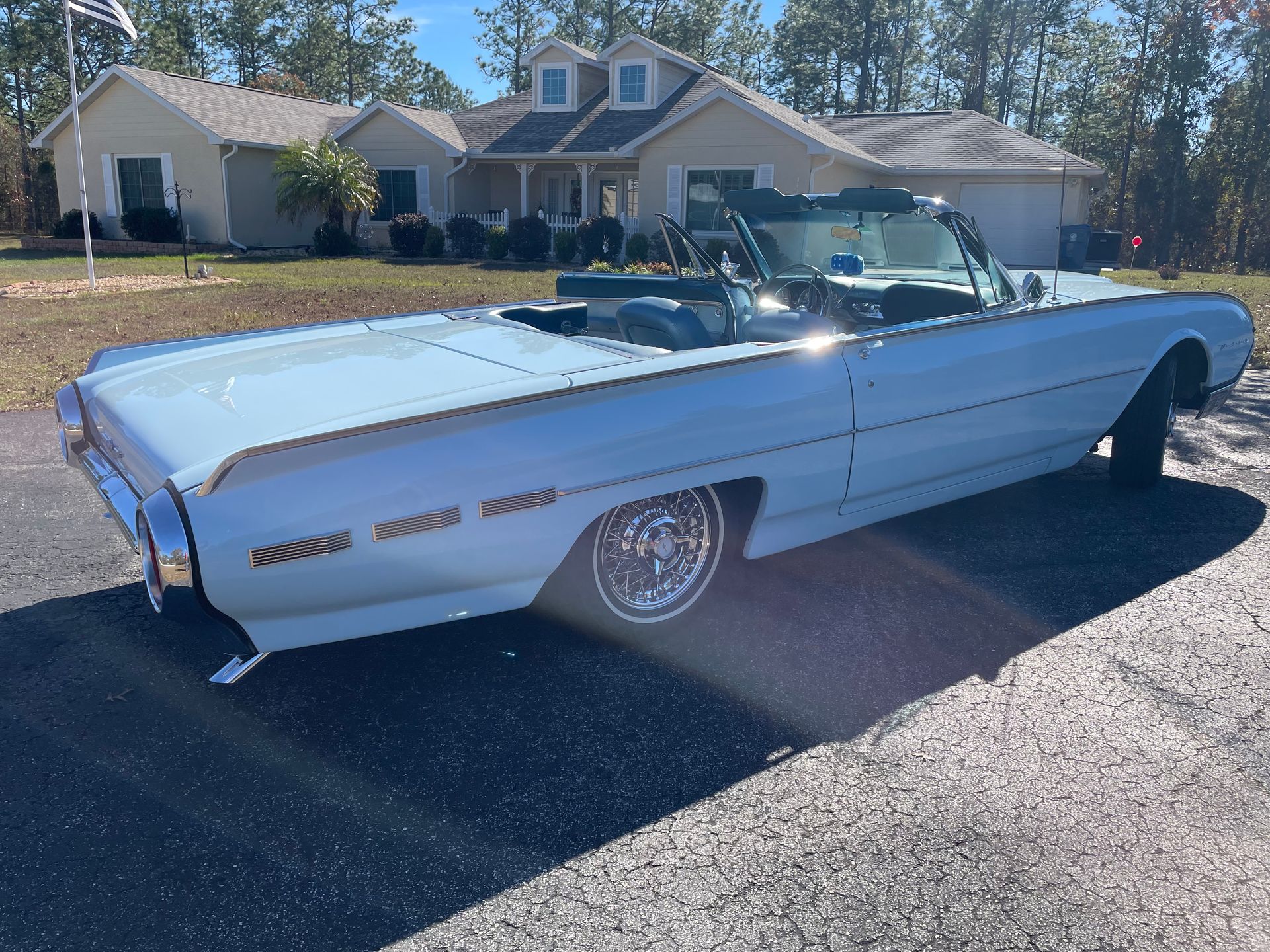 A light blue vintage Ford Thunderbird convertible parked on a gravel driveway in front of a suburban house.