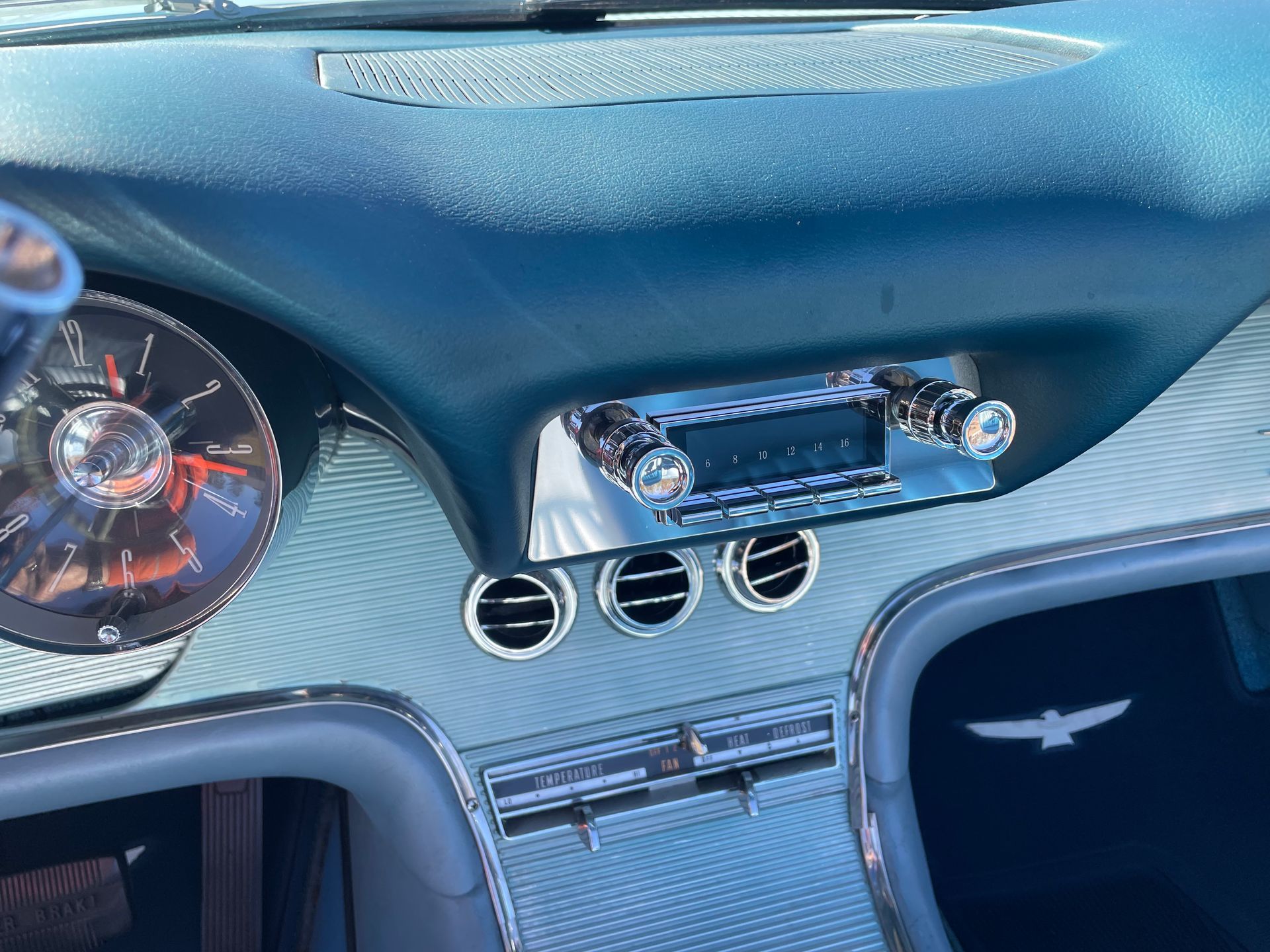 A close-up of a teal and metallic car dashboard featuring a chrome radio, circular air vents, and a Thunderbird emblem.