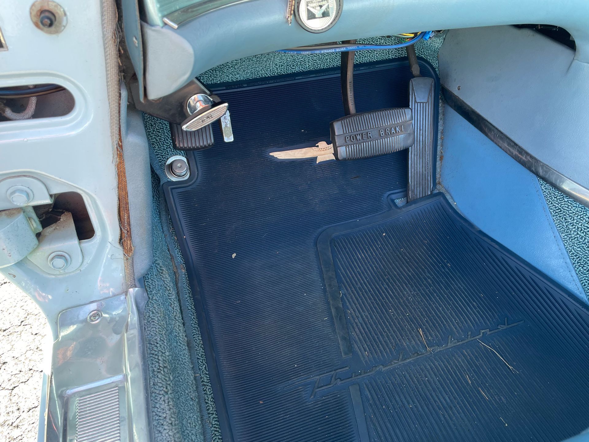The footwell of a vintage car with blue interior trim, a black ribbed floor mat, and a silver-pedal parking brake.