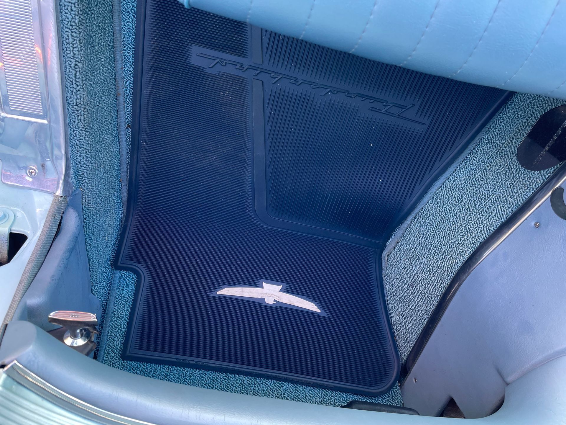 A blue car floor mat with a white Thunderbird emblem, placed on speckled blue carpeting inside a vehicle.