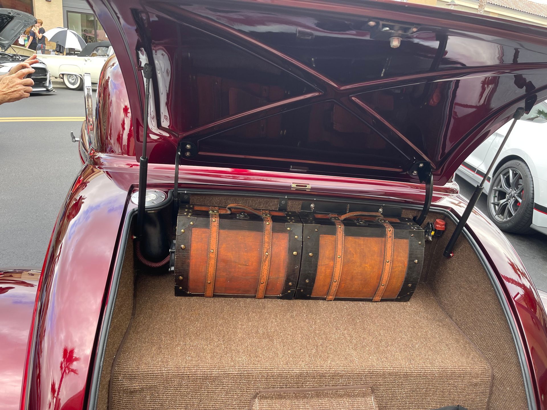 Open trunk of a deep red classic car with two leather vintage-style trunks inside and tan carpet lining.