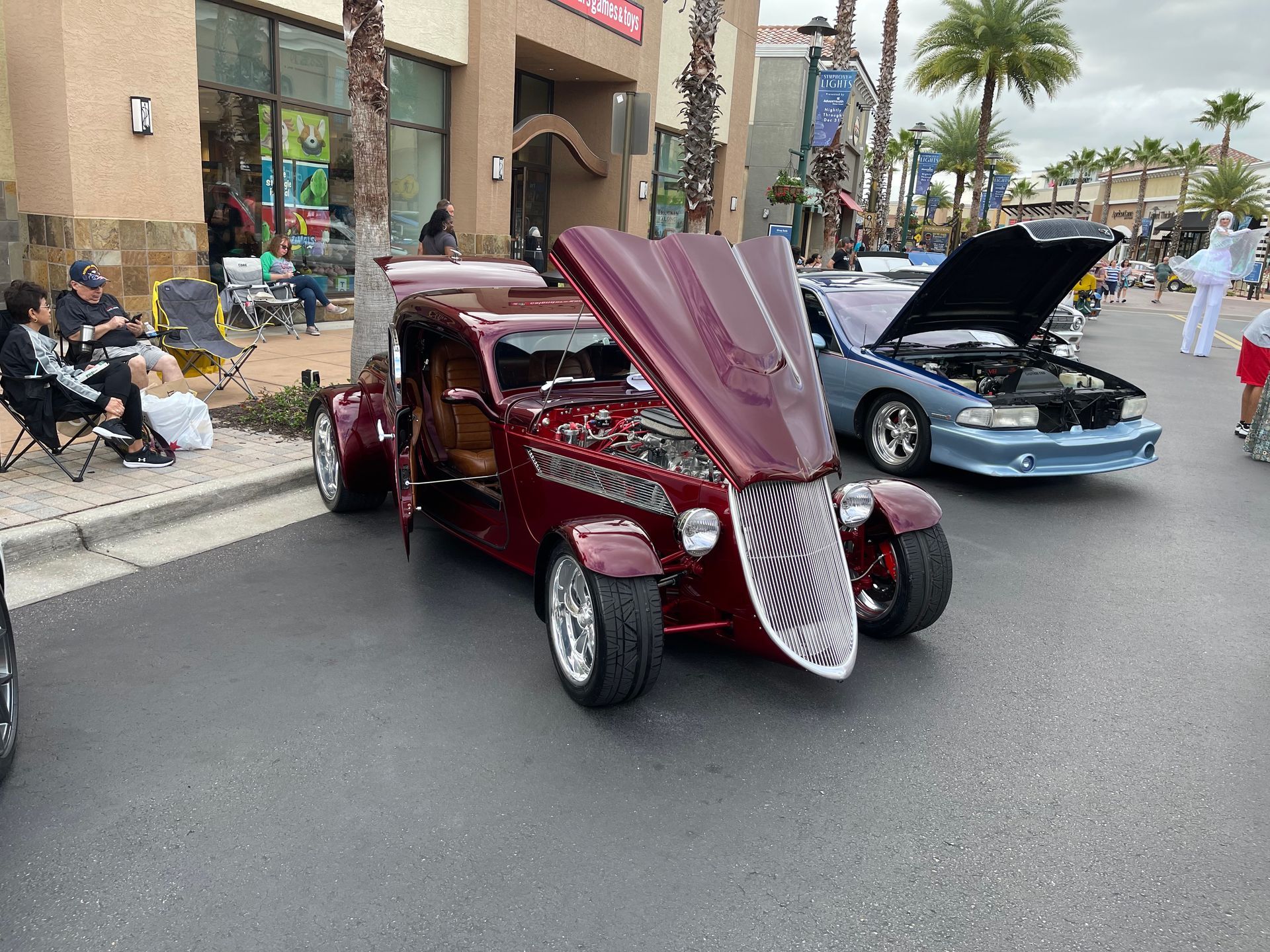 A maroon custom hot rod with an open hood sits parked next to a light blue car at an outdoor car show.