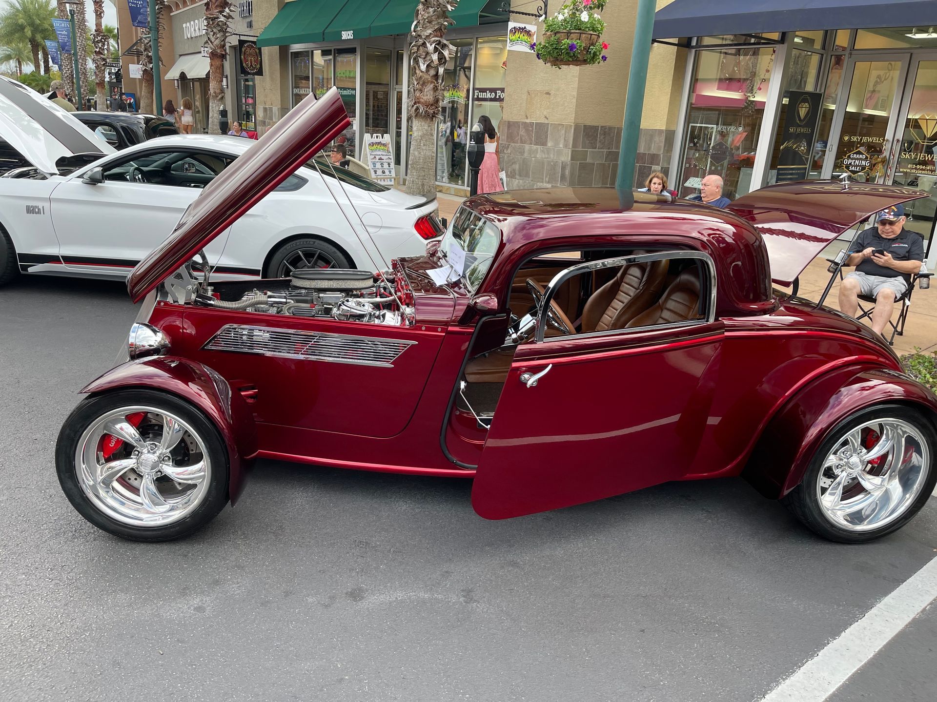 A deep red, modified classic hot rod with its hood and door open is parked outdoors on an asphalt lot.