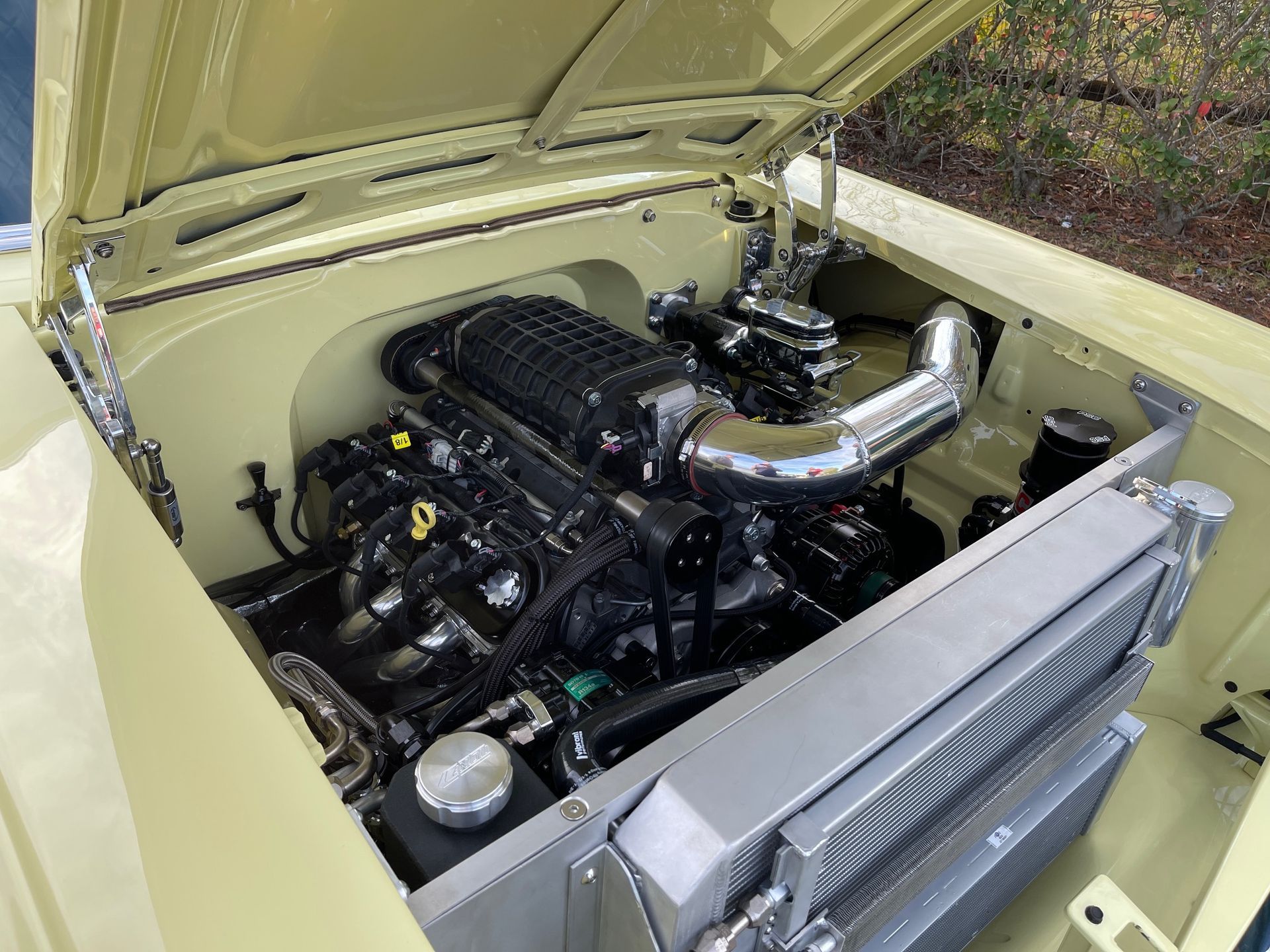 Open hood of a light yellow vintage car, showcasing a powerful black supercharged engine and a large aluminum radiator.