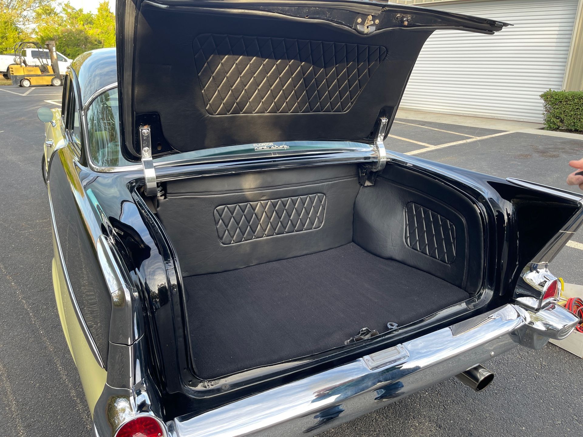 A black vintage car with an open trunk, featuring custom diamond-stitched interior panels, parked on an asphalt lot.