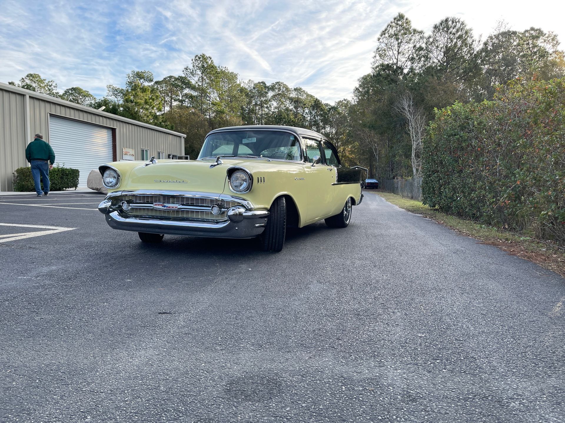 A pale yellow 1957 Chevrolet Bel Air parked on a paved lot beside a building and a hedge under a cloudy sky.