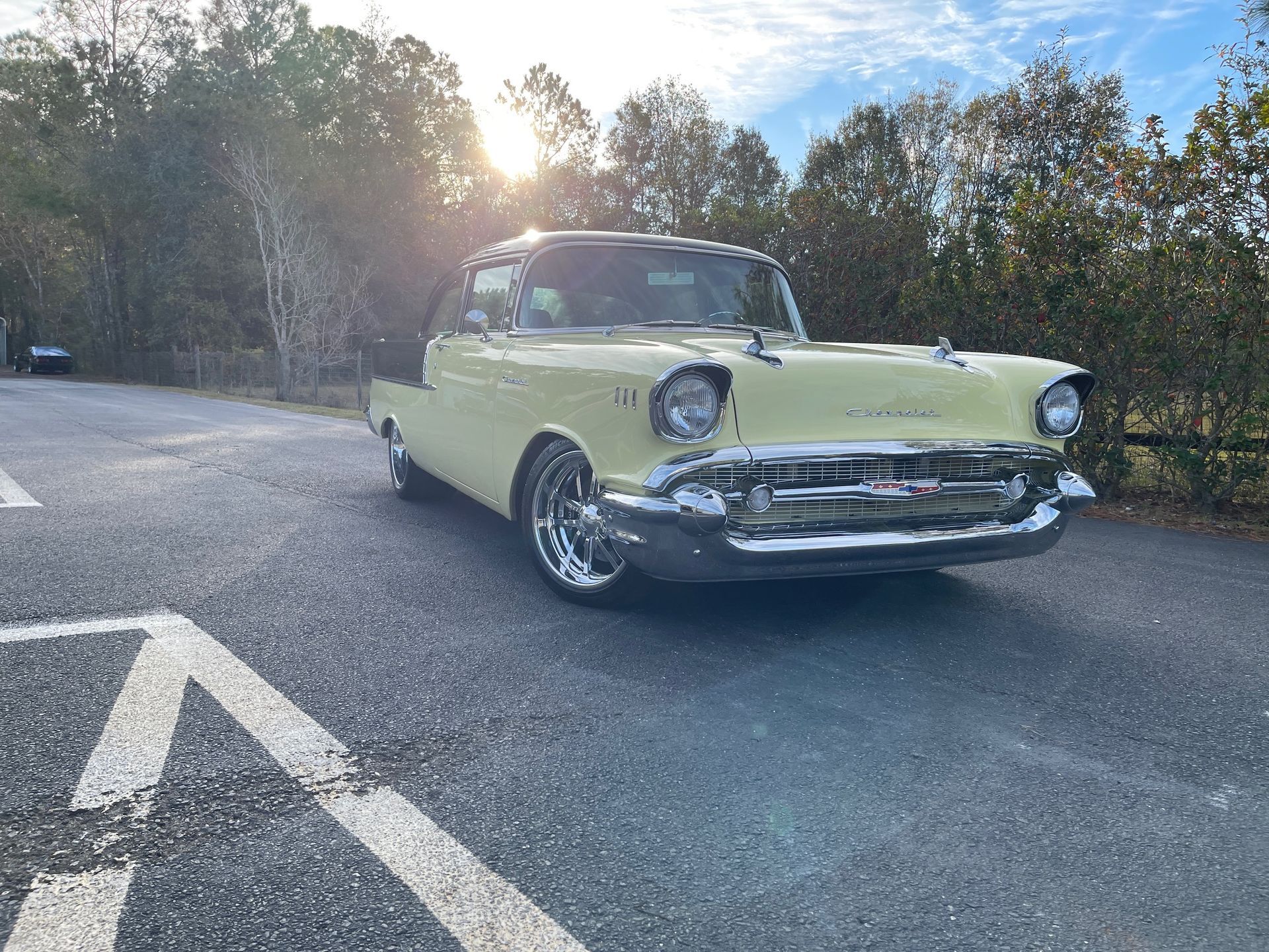 A pale yellow vintage Chevrolet parked on an asphalt road with trees in the background under a bright, sunny sky.