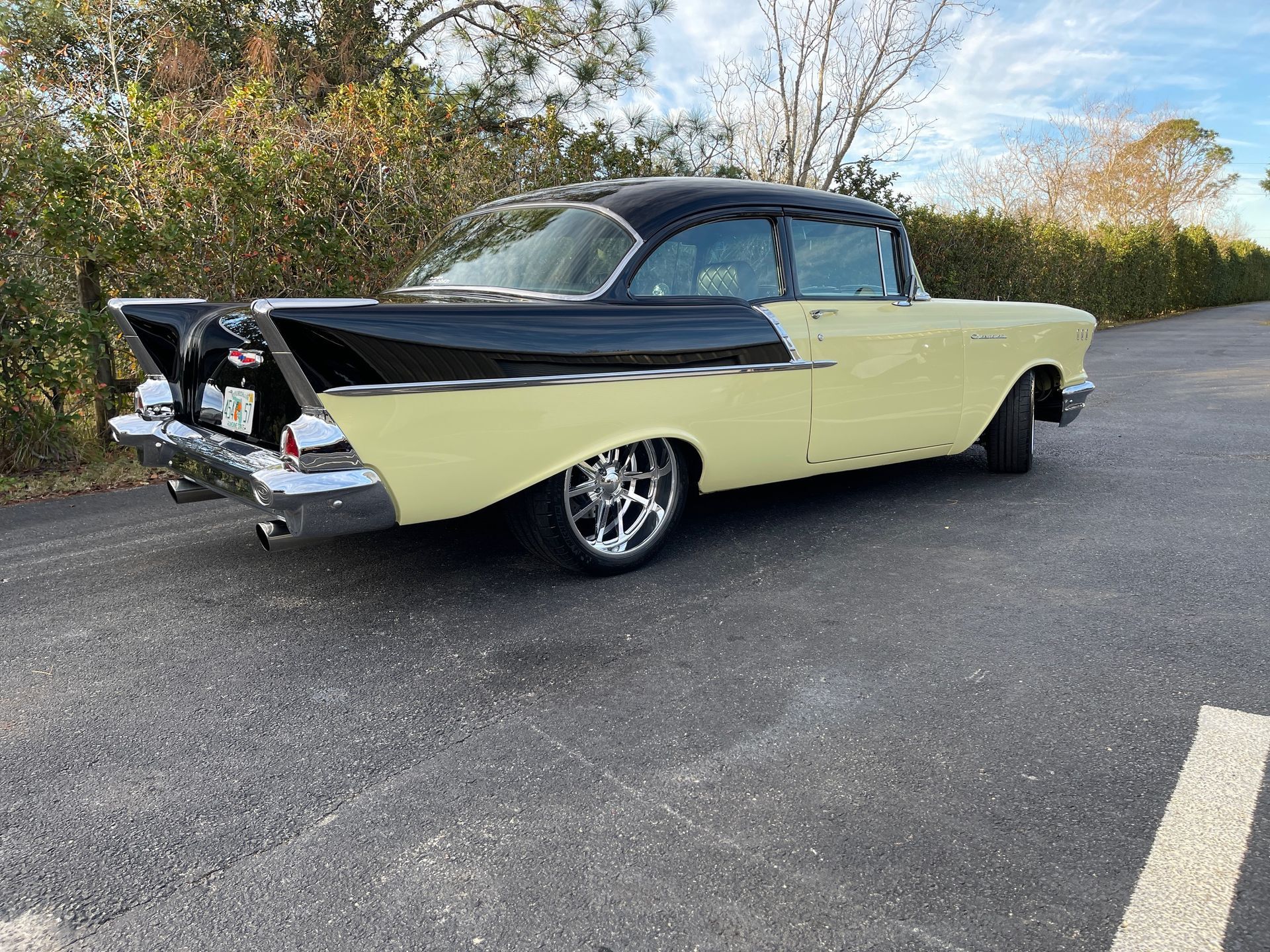 A vintage two-tone black and pale yellow 1957 Chevrolet parked on an asphalt road next to greenery.