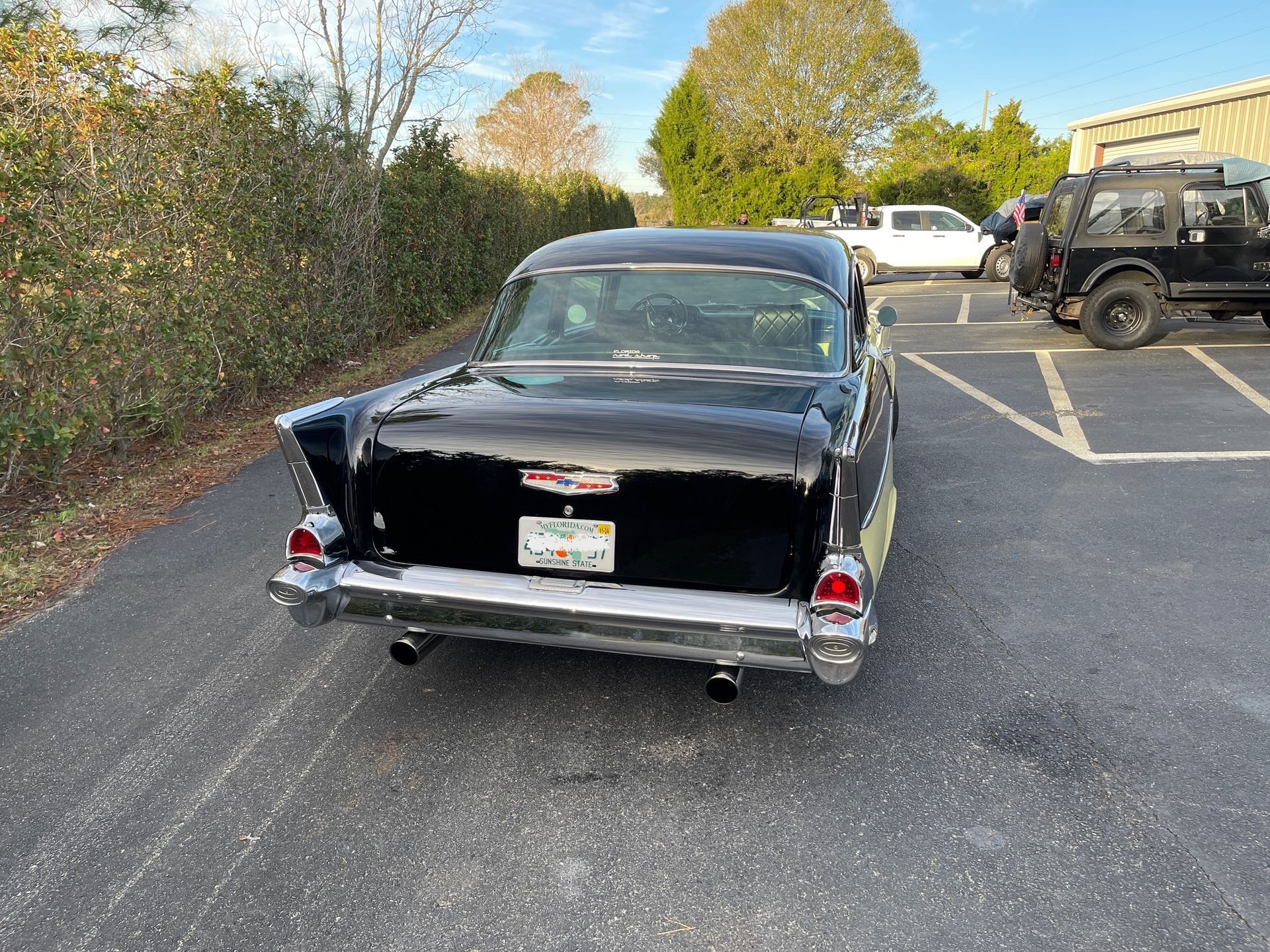 A black vintage 1957 Chevrolet Bel Air parked in an asphalt lot with other vehicles visible in the background.