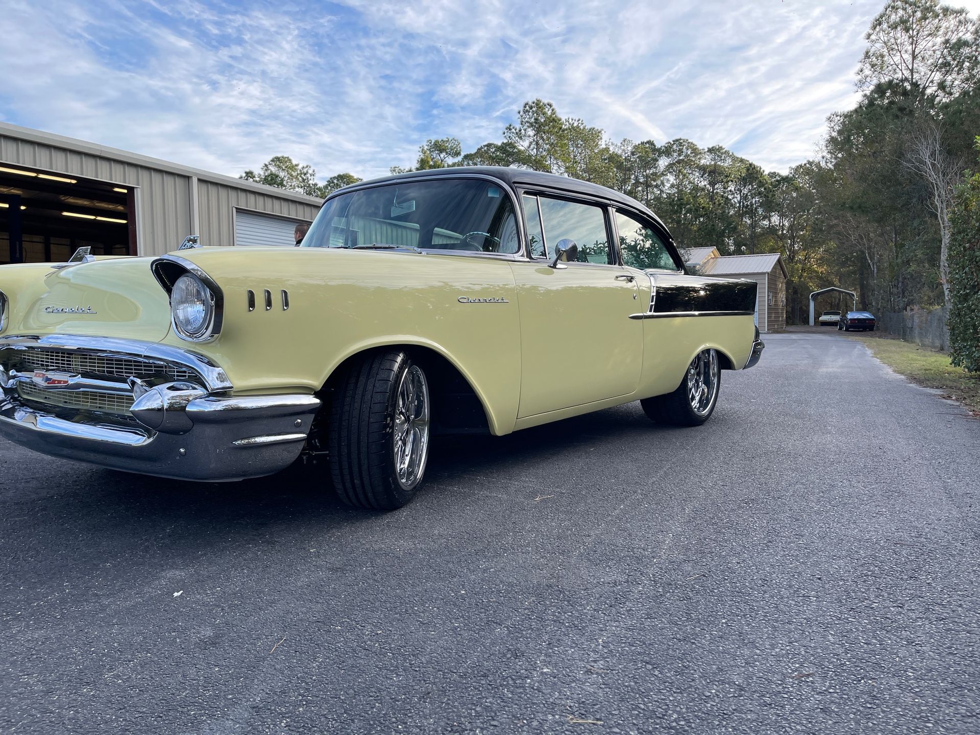 A pale yellow and black 1957 Chevrolet Bel Air parked on a gravel driveway outdoors under a partly cloudy sky.