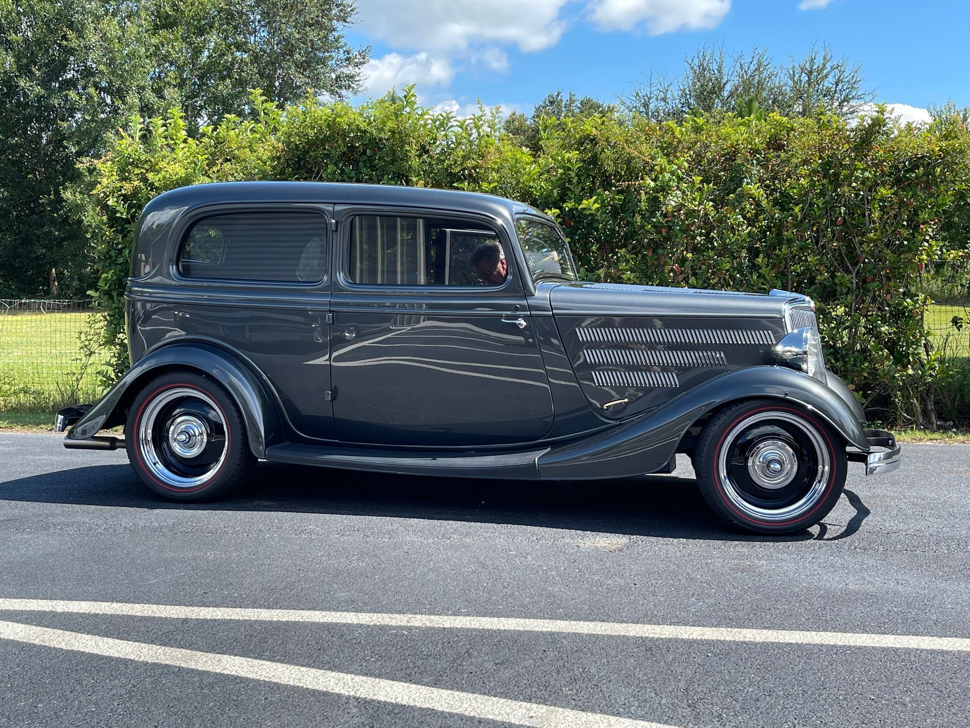 A dark grey vintage sedan parked on an asphalt road in front of a green hedge on a sunny day.