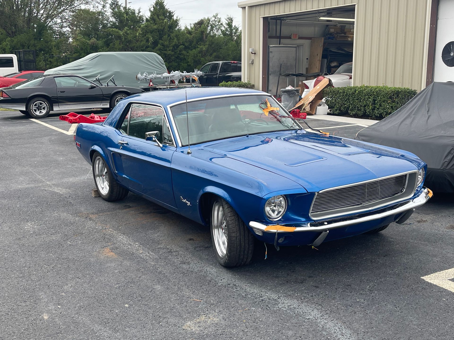 A bright blue, vintage Ford Mustang parked in an outdoor lot in front of a garage building.