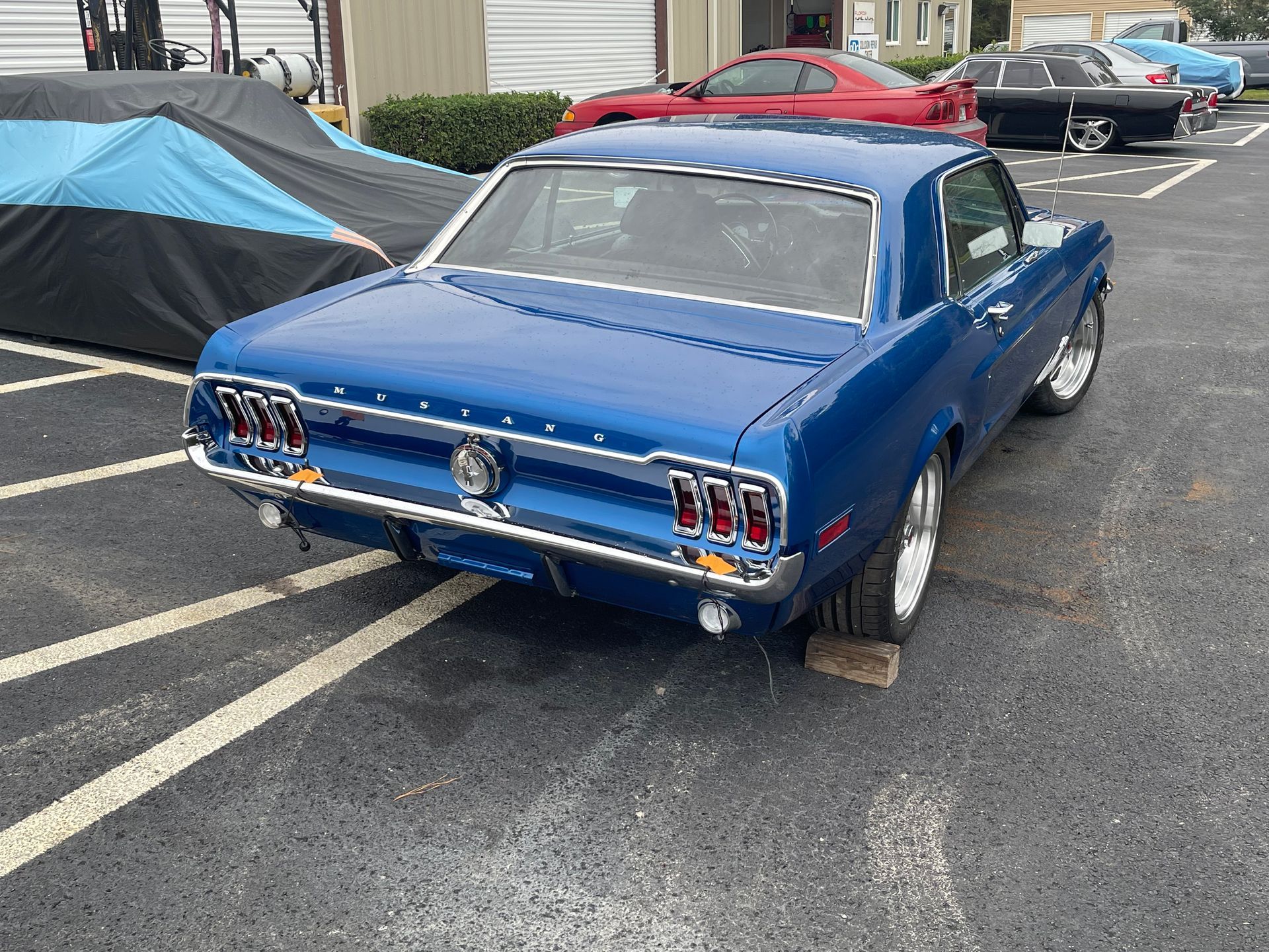 A blue 1968 Ford Mustang coupe parked in an asphalt parking lot, next to a car covered by a blue and black tarp.