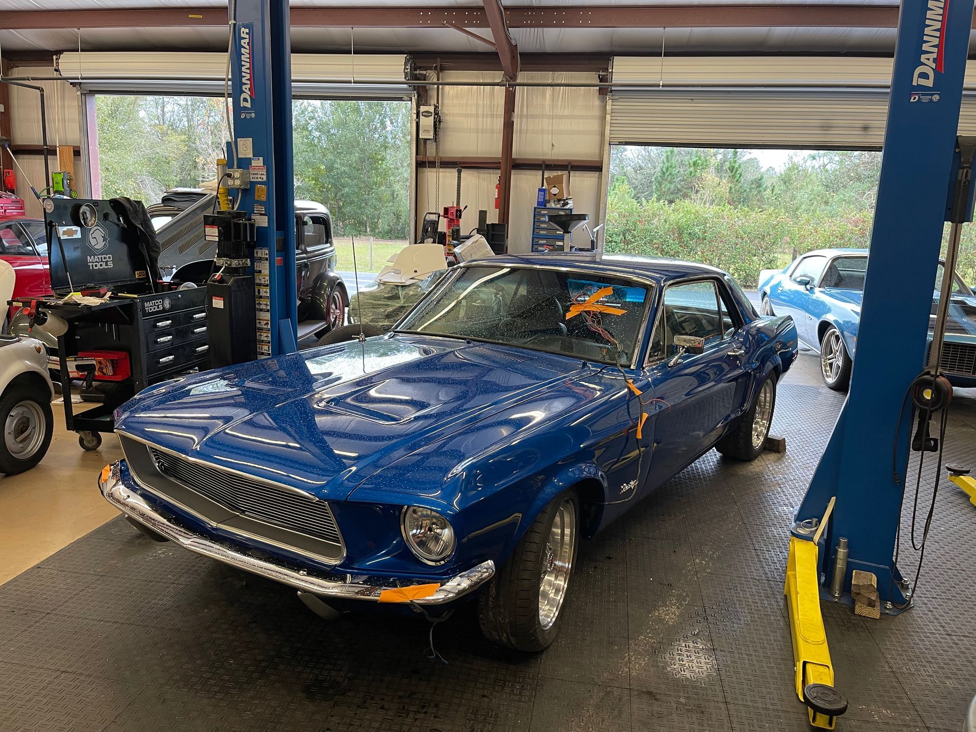 A blue vintage Mustang parked inside an auto repair shop between the posts of a car lift.