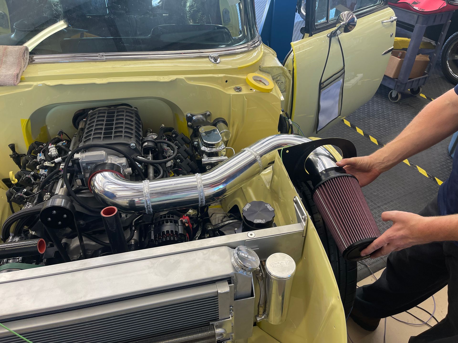 A pair of hands holds a cylindrical air filter next to the intake piping of an engine in a yellow classic car.