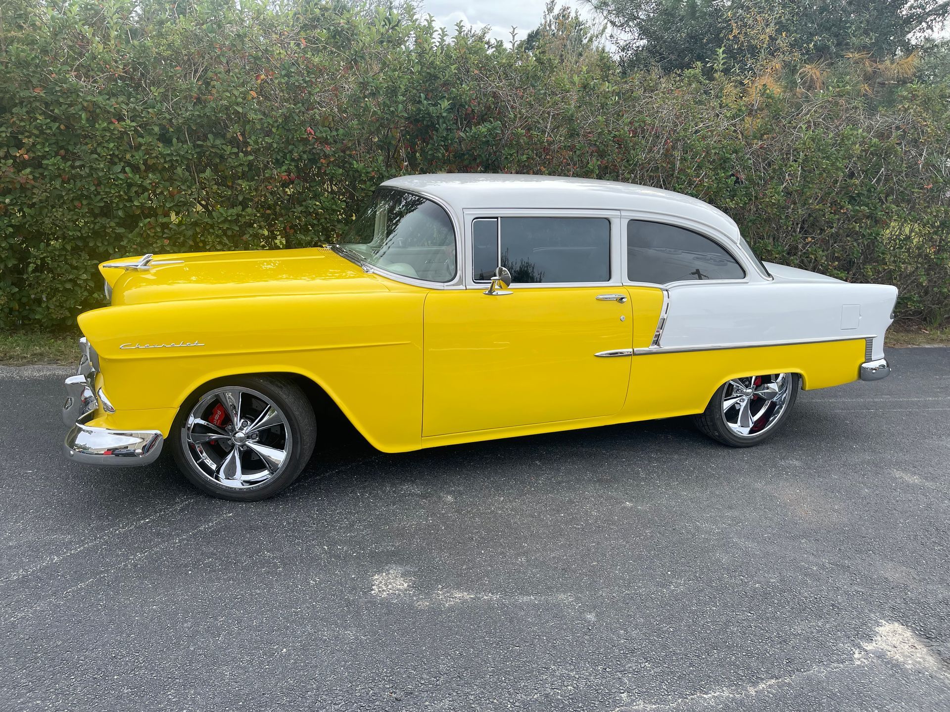 A yellow and white two-tone 1955 Chevrolet classic car parked on asphalt in front of a hedge.