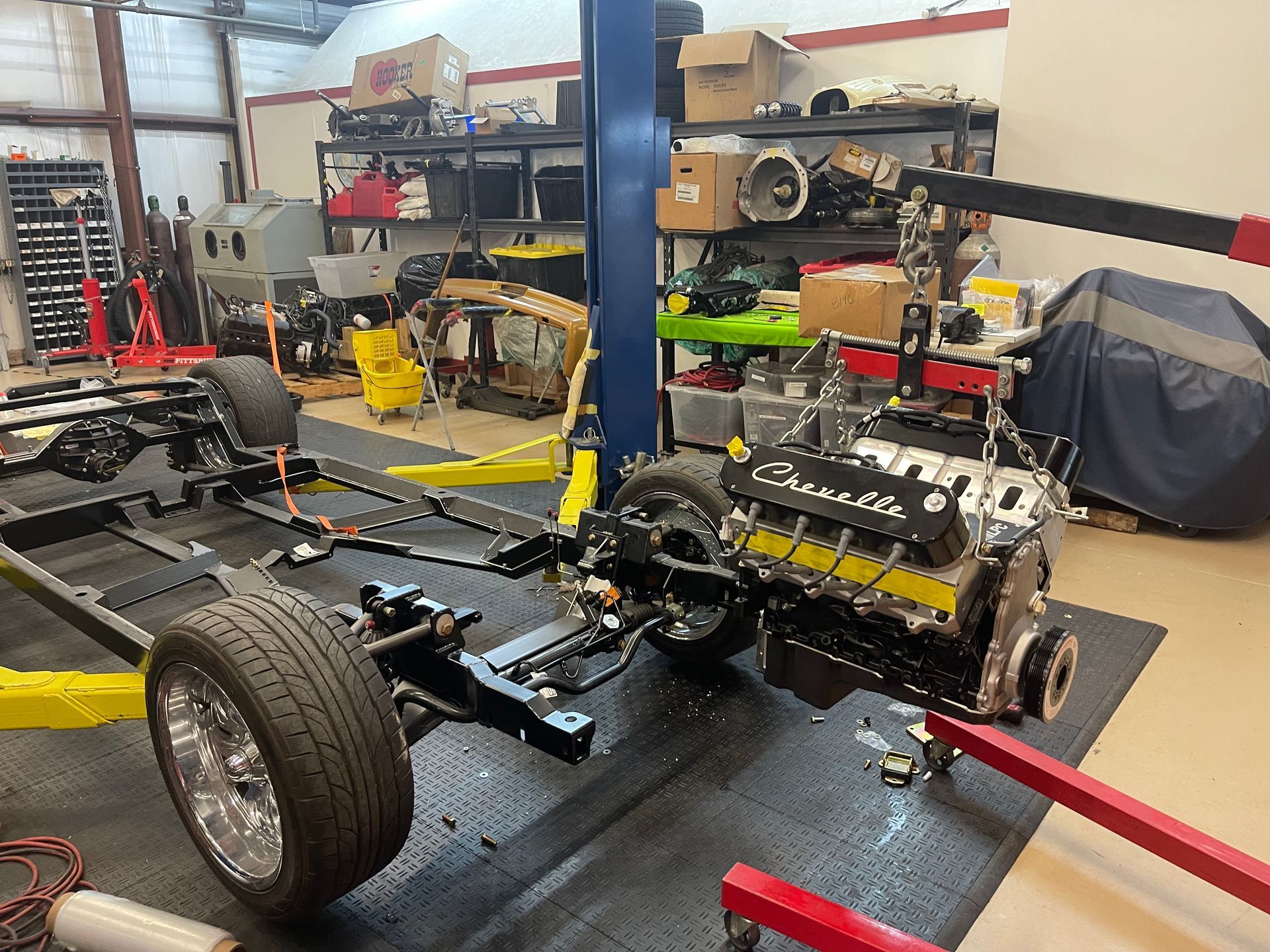 An engine with black Chevrolet valve covers hangs from a red crane over a vehicle chassis in a garage.