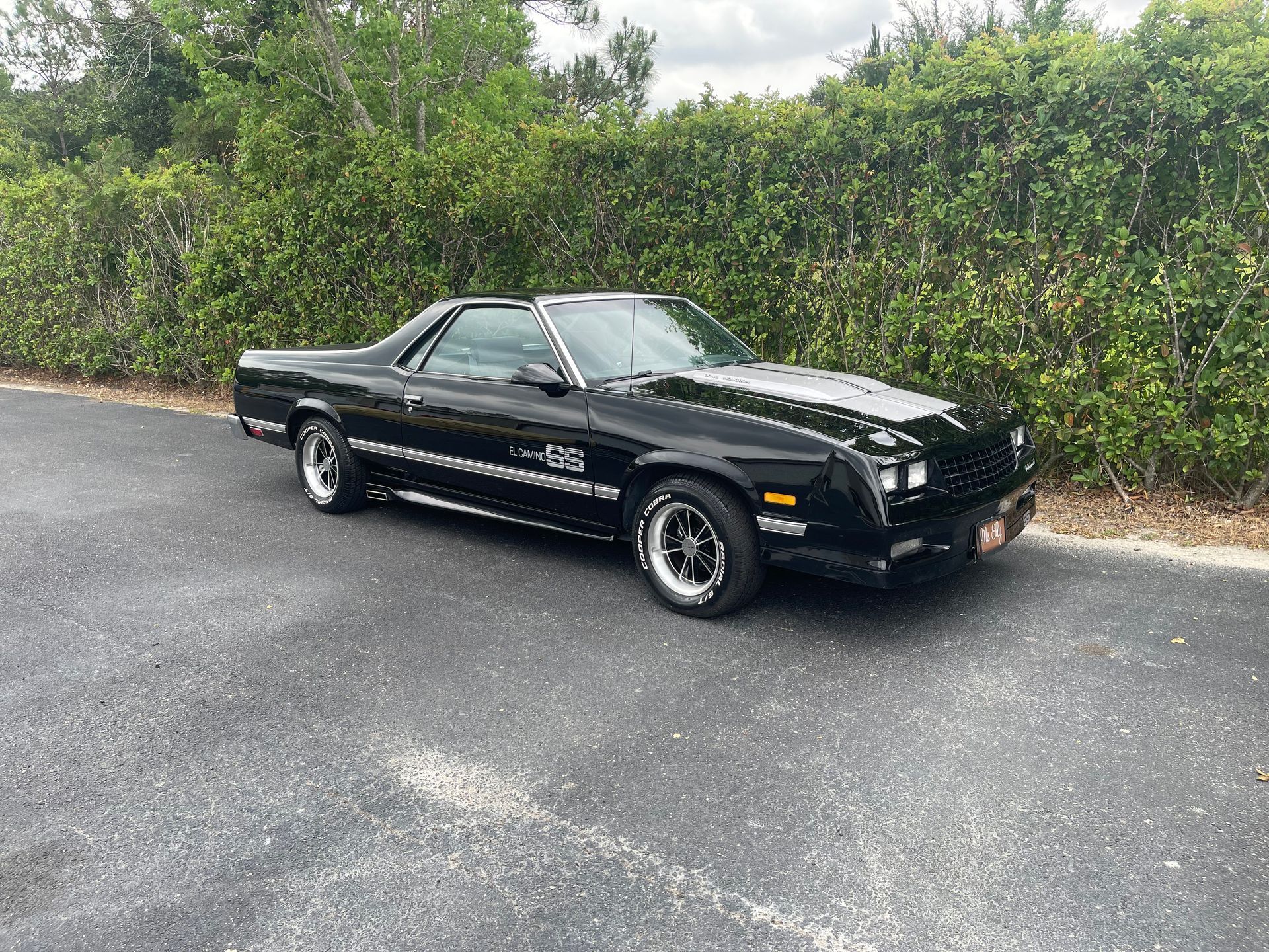A black Chevrolet El Camino parked on an asphalt driveway in front of a green hedge.