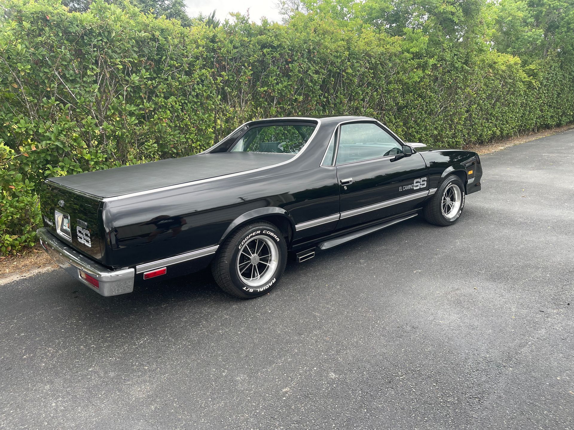 A black Chevrolet El Camino parked on an asphalt driveway next to a tall green hedge.