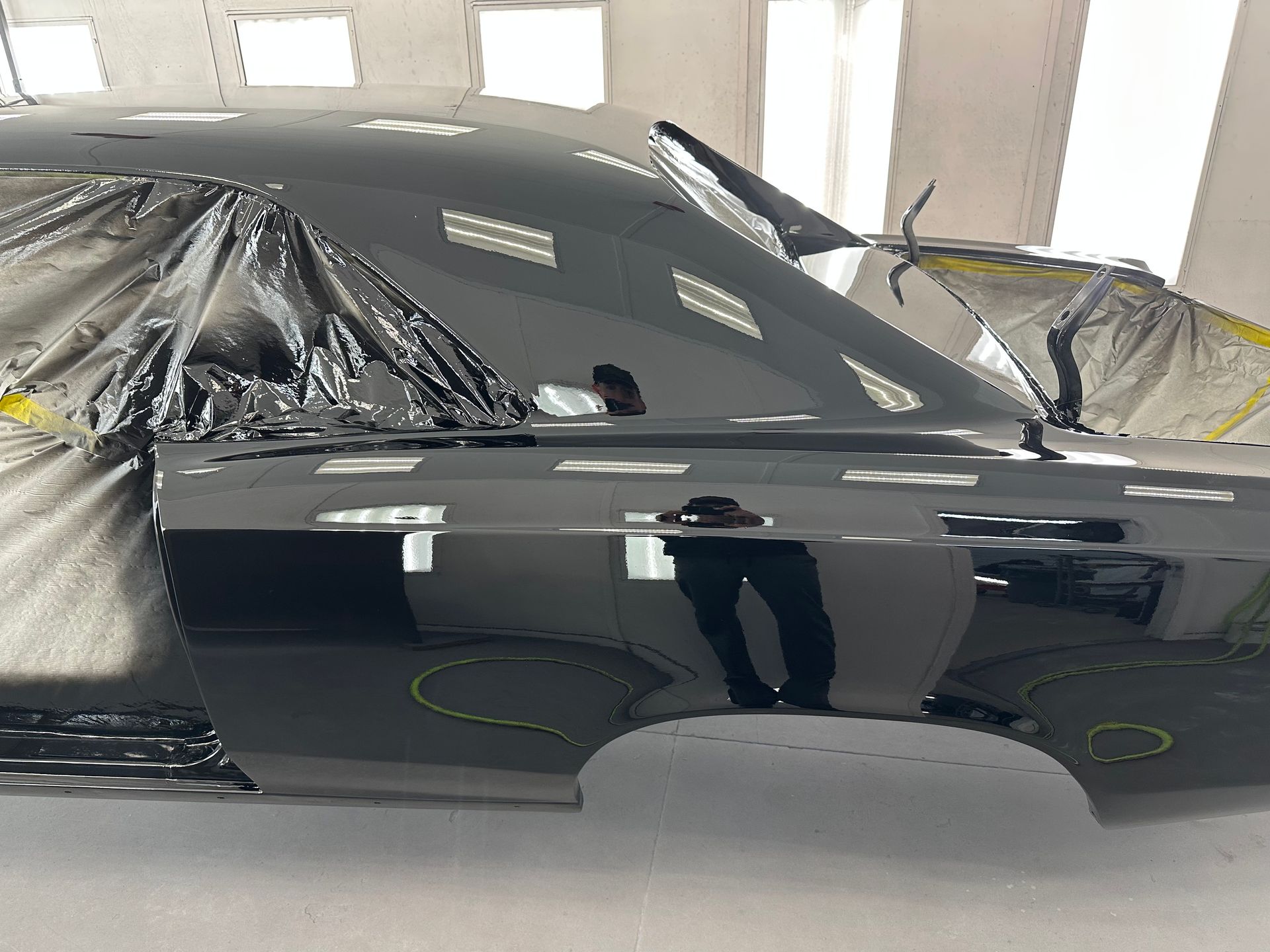 A shiny, black-painted car body section inside a professional spray booth with masking plastic on the windows.