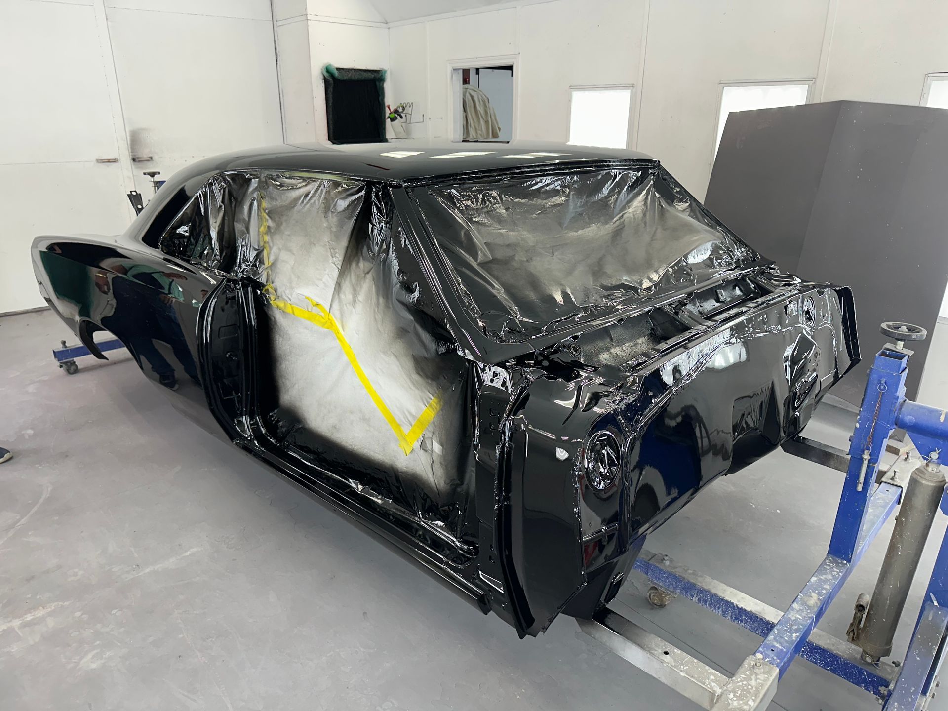 A black car body on a rotisserie stand in a workshop, with the windows masked off for painting.