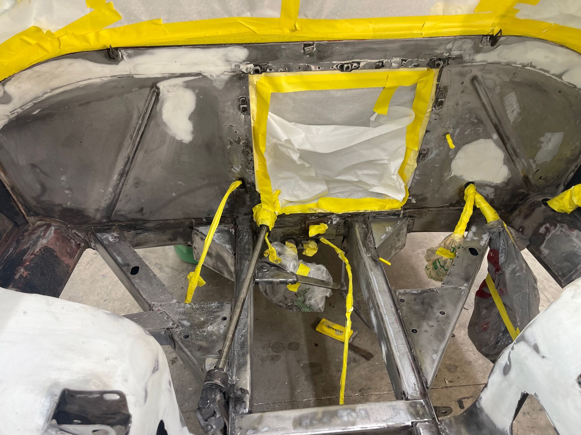 A partially disassembled vehicle frame with metal panels, masked with yellow tape and white plastic sheets for painting.
