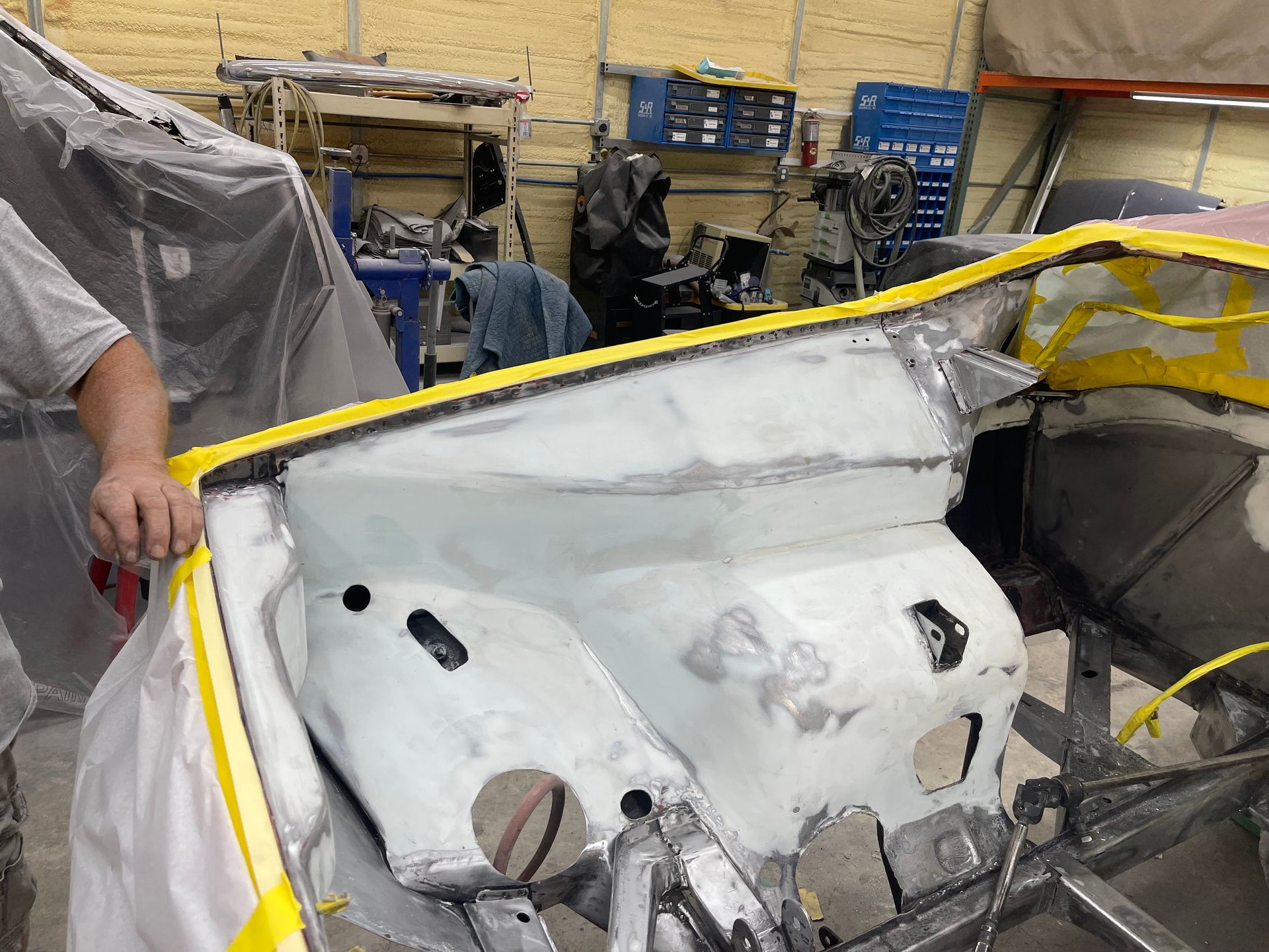 A person works on a grey, sanded engine bay of a vehicle in an auto body shop, bordered by yellow masking tape.