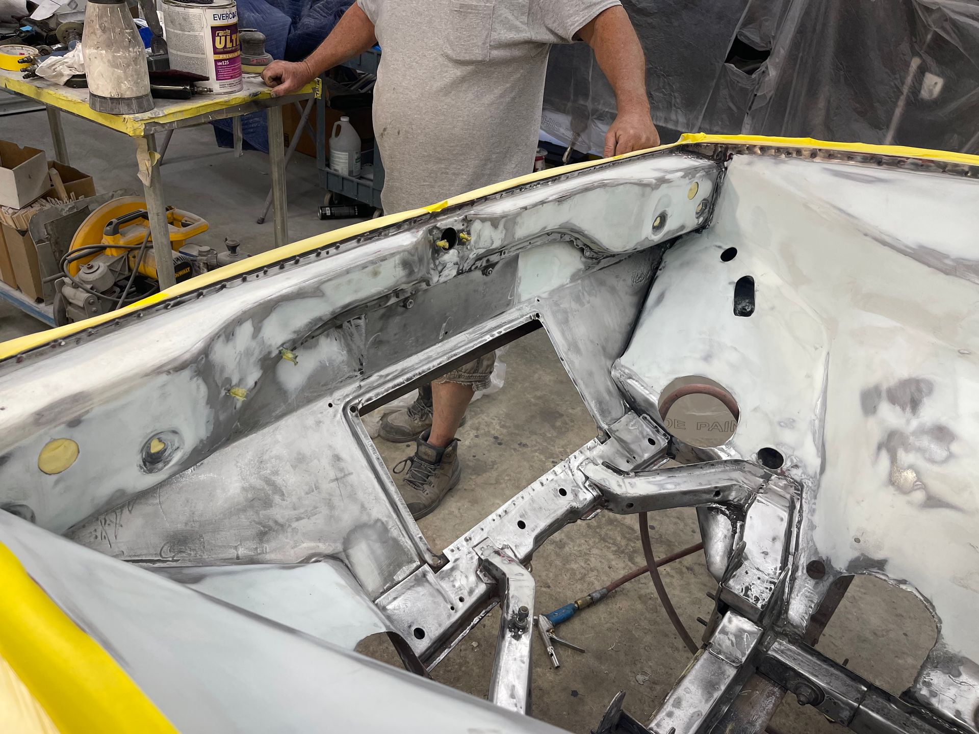 A person stands by a car engine bay under restoration, showing the bare metal frame and inner firewall in a garage.